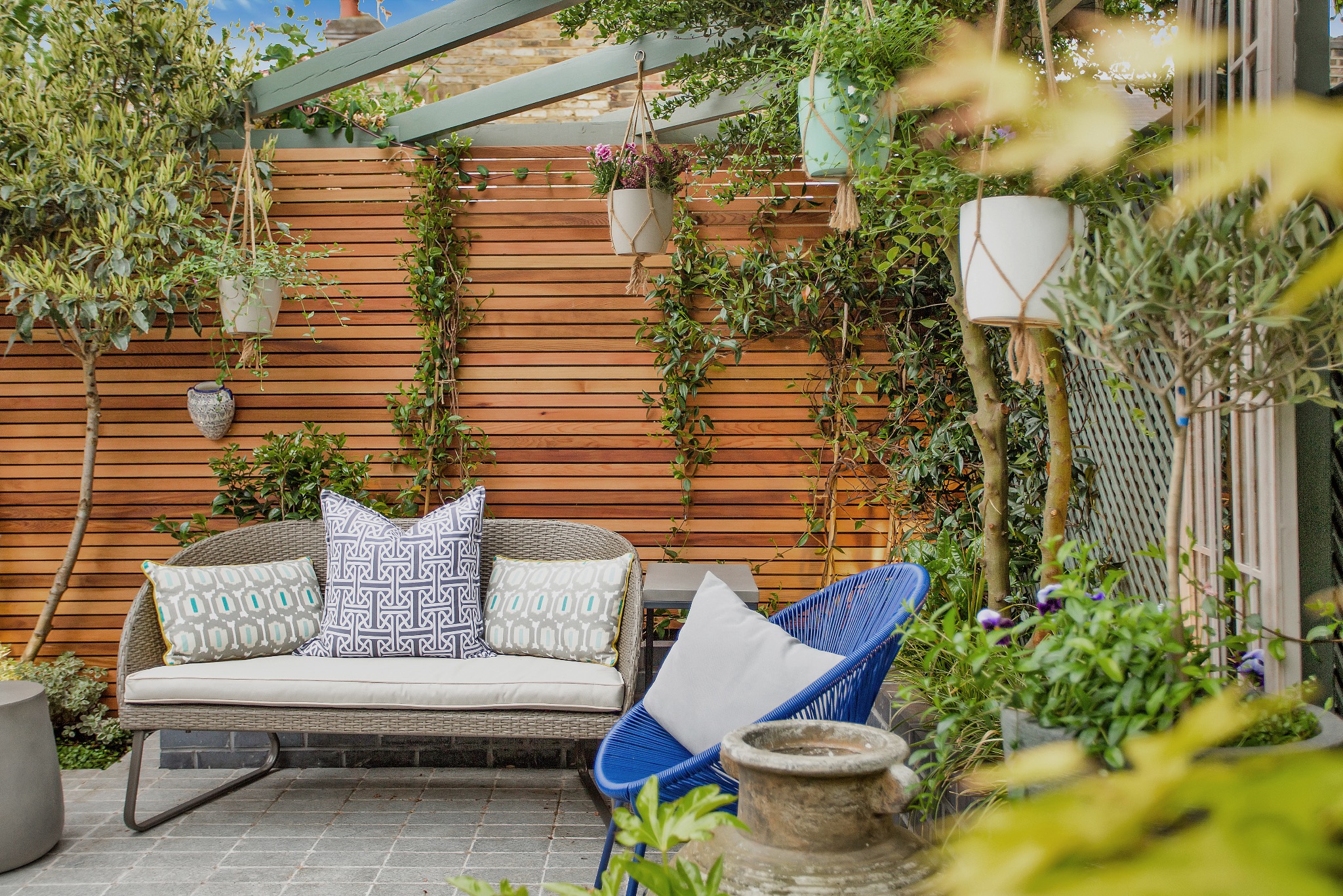 Aranżacja balkonu z drewnianą ścianą, wiklinową sofą z wzorzystymi poduszkami, niebieskim krzesłem z poduszką, wiszącymi doniczkami i roślinnością.