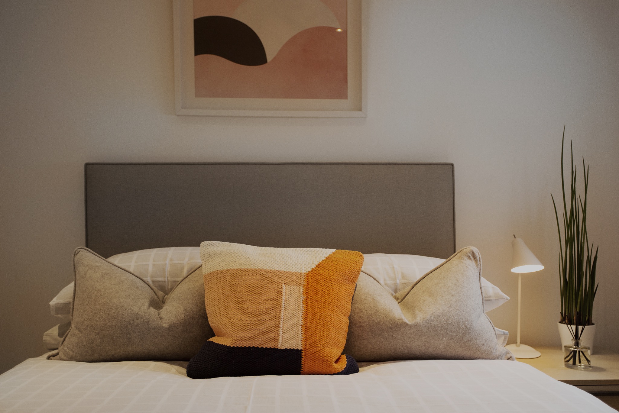 Minimalistyczna sypialnia z geometryczną poduszką w odcieniach beżu i pomarańczy na łóżku z szarą ramą, nad łóżkiem abstrakcyjny obraz w pastelowych kolorach, obok lampka nocna i roślina w szklanym...