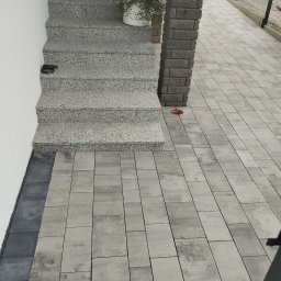 Usługi budowlane - Ułożona szara kostka brukowa na podjeździe i schody z tego samego materiału, z donicą z roślinami na górnym stopniu i ceglanym słupkiem obok.