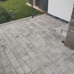 Usługi budowlane - Świeżo ułożona, szara kostka brukowa tworząca taras przed domem z nowoczesnym ogrodzeniem i fragmentem trawnika w tle.