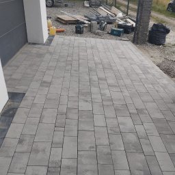 Usługi budowlane - Nowo ułożona, szara kostka brukowa przed budynkiem z widocznymi narzędziami i materiałami budowlanymi w tle.