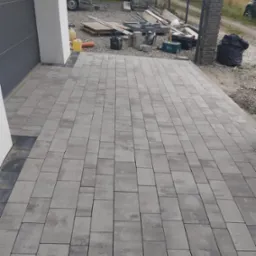 Nowo ułożona, szara kostka brukowa przed budynkiem z widocznymi narzędziami i materiałami budowlanymi w tle.