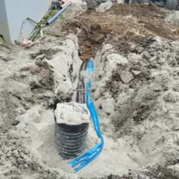 Wykop z rurami i studzienką kanalizacyjną, częściowo zasypany piaskiem, z widocznym wykopem i koparką w tle.