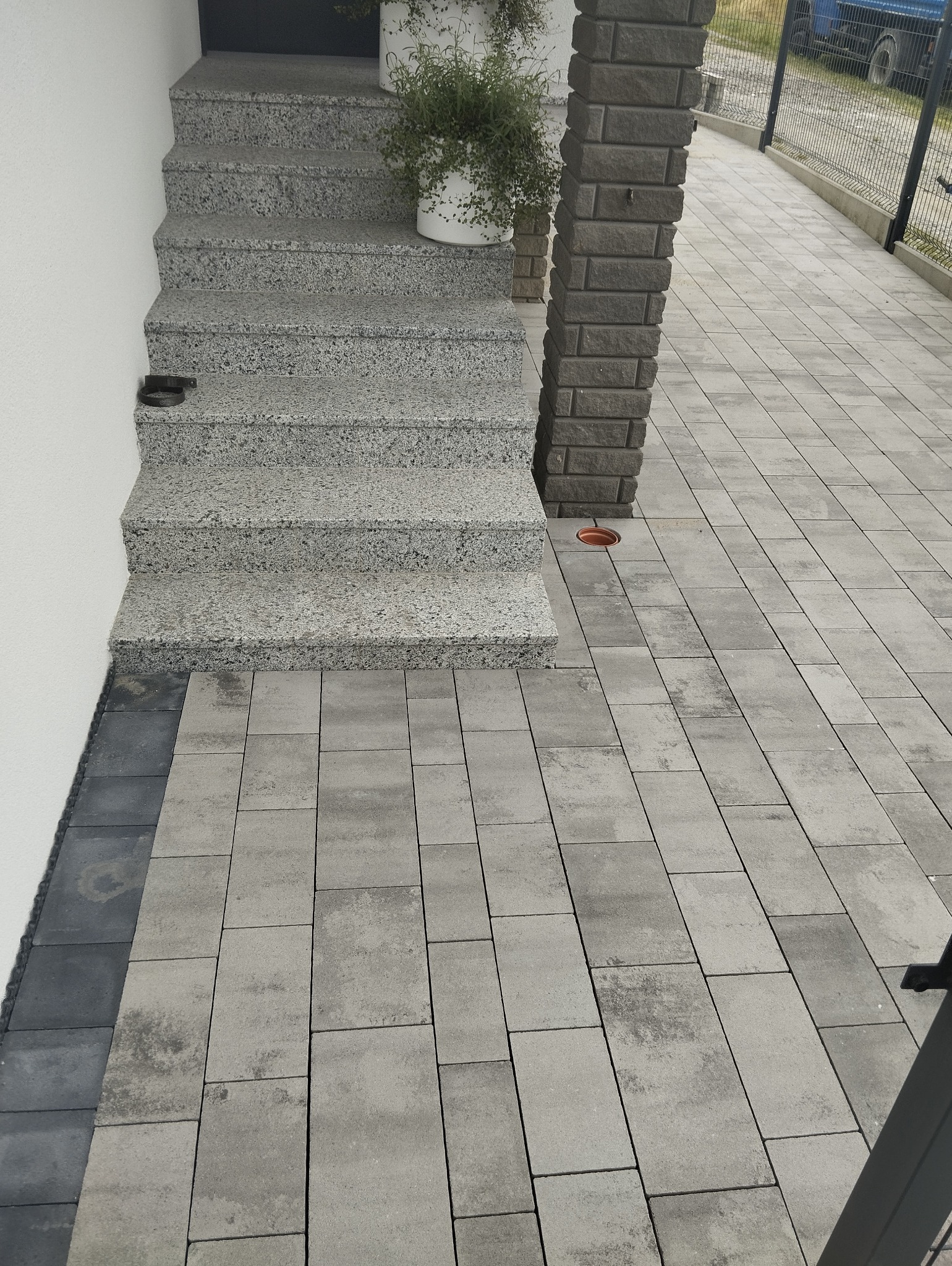 Ułożona szara kostka brukowa na podjeździe i schody z tego samego materiału, z donicą z roślinami na górnym stopniu i ceglanym słupkiem obok.
