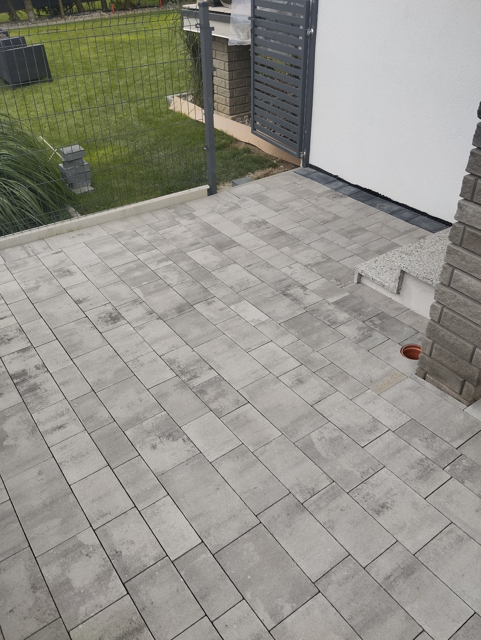 Świeżo ułożona, szara kostka brukowa tworząca taras przed domem z nowoczesnym ogrodzeniem i fragmentem trawnika w tle.
