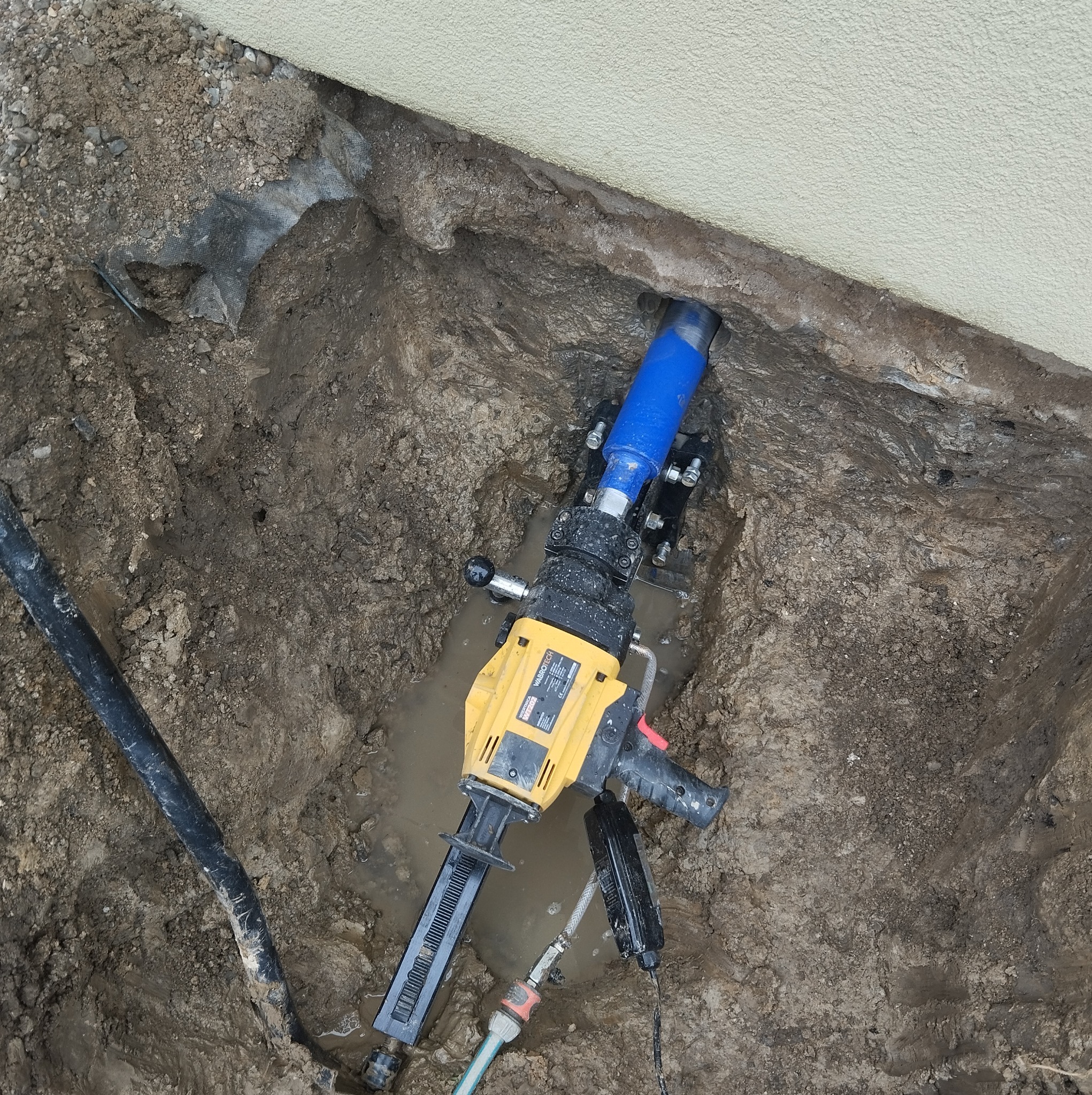Wiertnica diamentowa WABROTECH WT200 w wykopie, wiercenie rdzeniowe w pobliżu fundamentu budynku, widoczny fragment niebieskiego przewodu, woda w wykopie.