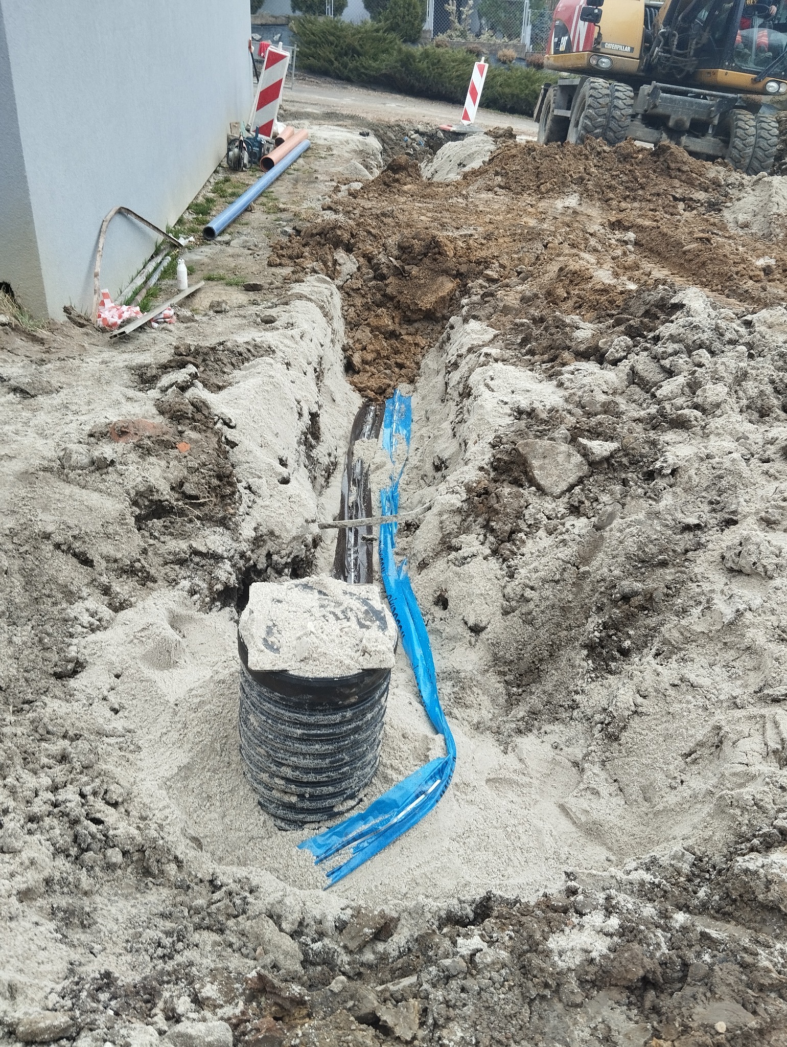 Wykop z rurami i studzienką kanalizacyjną, częściowo zasypany piaskiem, z widocznym wykopem i koparką w tle.
