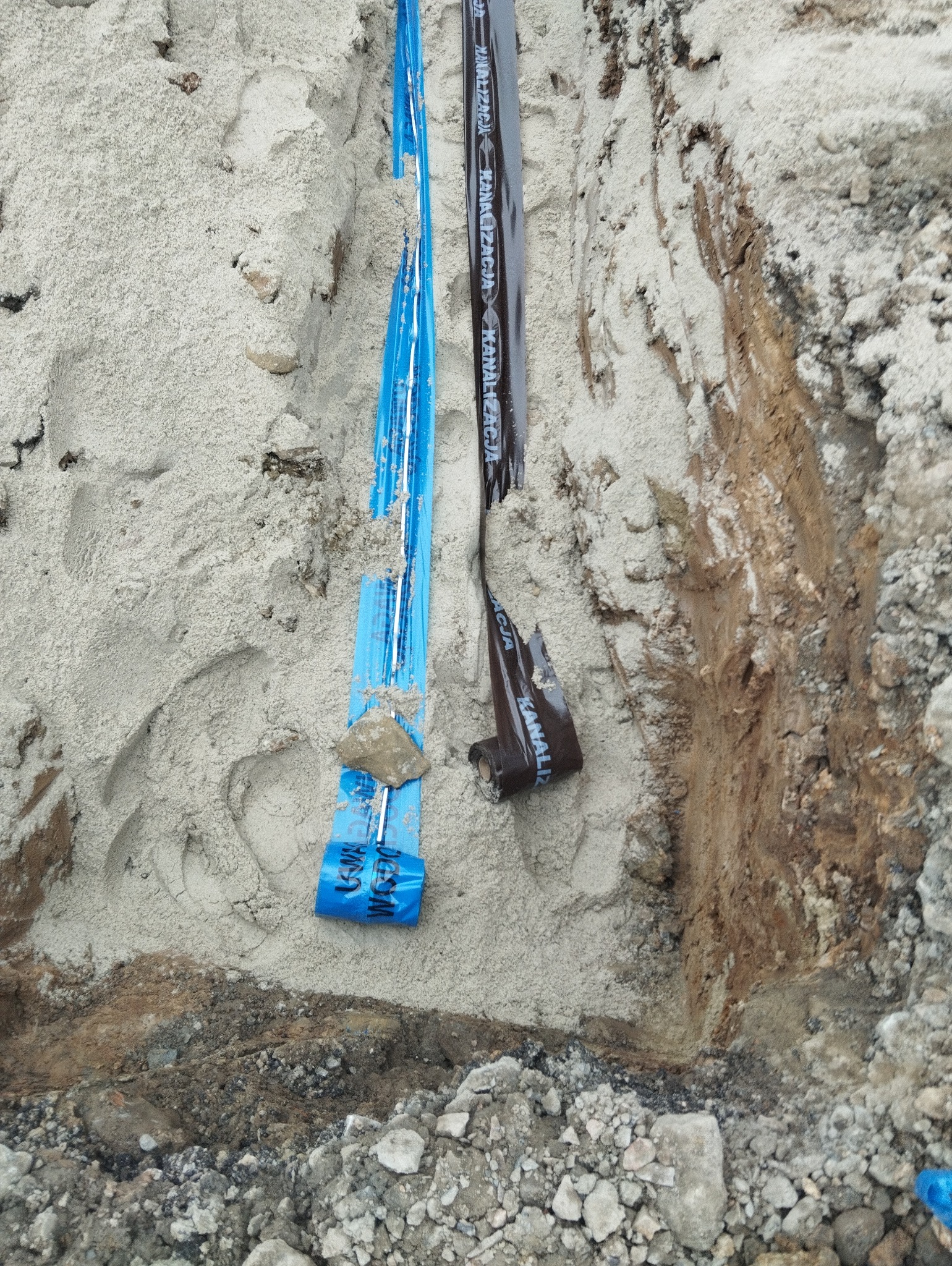 Wykop w ziemi z widocznymi rurami oznaczonymi taśmami ostrzegawczymi w kolorze niebieskim i brązowym z napisem 'Kanalizacja'.