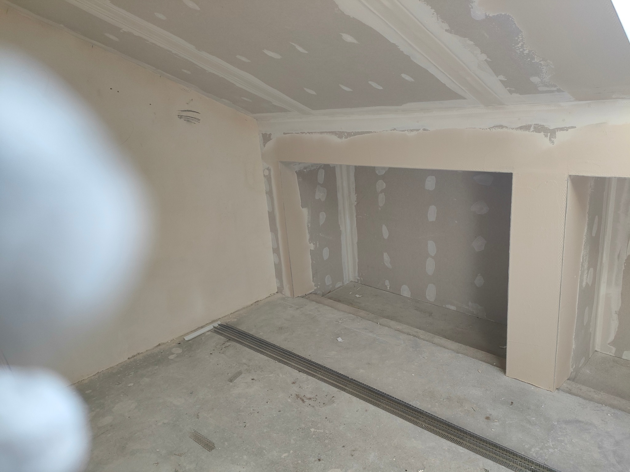 Wnętrze w trakcie prac wykończeniowych, widoczne ściany i sufit po szpachlowaniu, przygotowane do dalszych prac, na podłodze listwy metalowe.