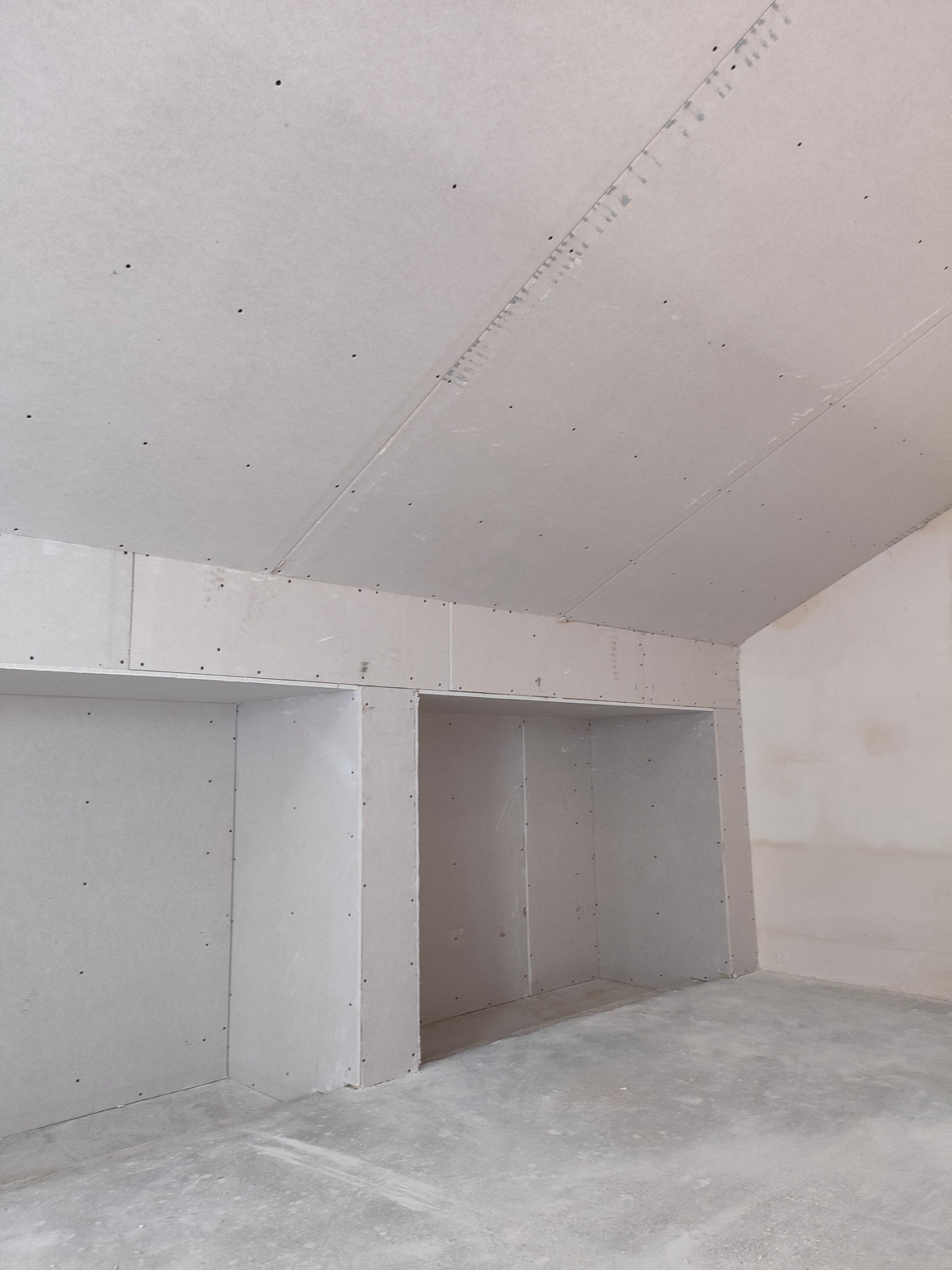 Pokój w trakcie wykańczania, widoczne ściany i sufit pokryte szarą płytą, przygotowane wnęki na szafy w zabudowie, szpachlowane łączenia płyt.