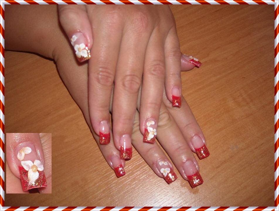 Dłonie z czerwonymi tipsami zdobionymi brokatem i trójwymiarowymi białymi kwiatkami, wykonane w salonie w Warszawie, na tle drewnianego blatu. Dodatkowe zbliżenie na jeden z paznokci prezentuje...