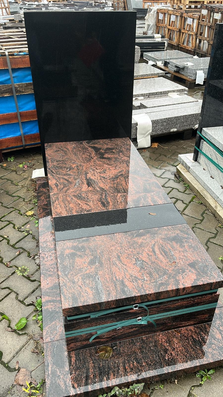 Czarny i czerwono-brązowy nagrobek z granitu na zewnątrz w Chojnicach. Widoczne palety z innymi elementami kamiennymi w tle.