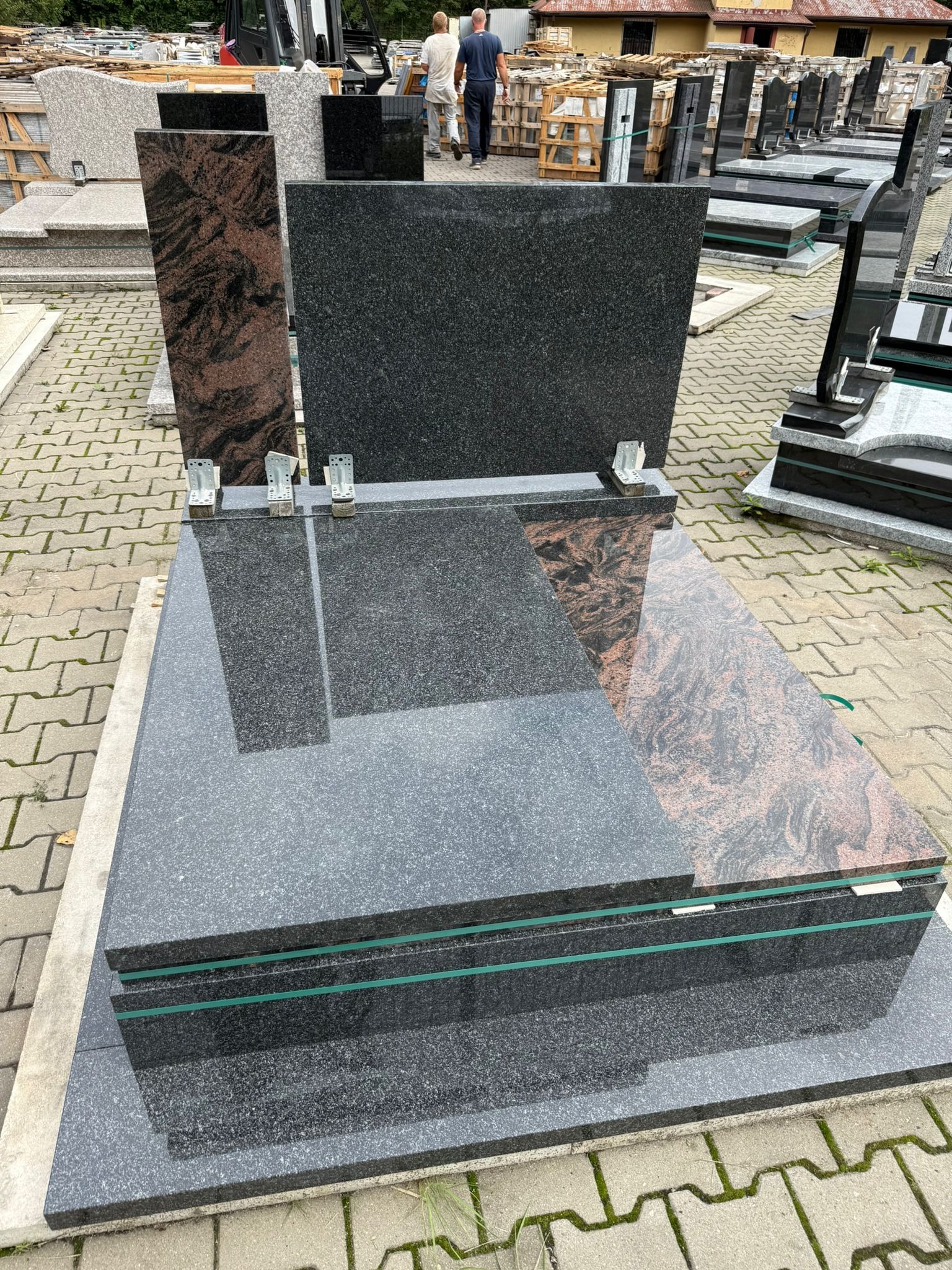 Wystawa nagrobków z granitu w różnych kolorach i wzorach, zabezpieczonych zieloną taśmą, na zewnątrz, z osobami w tle. Widok z góry.