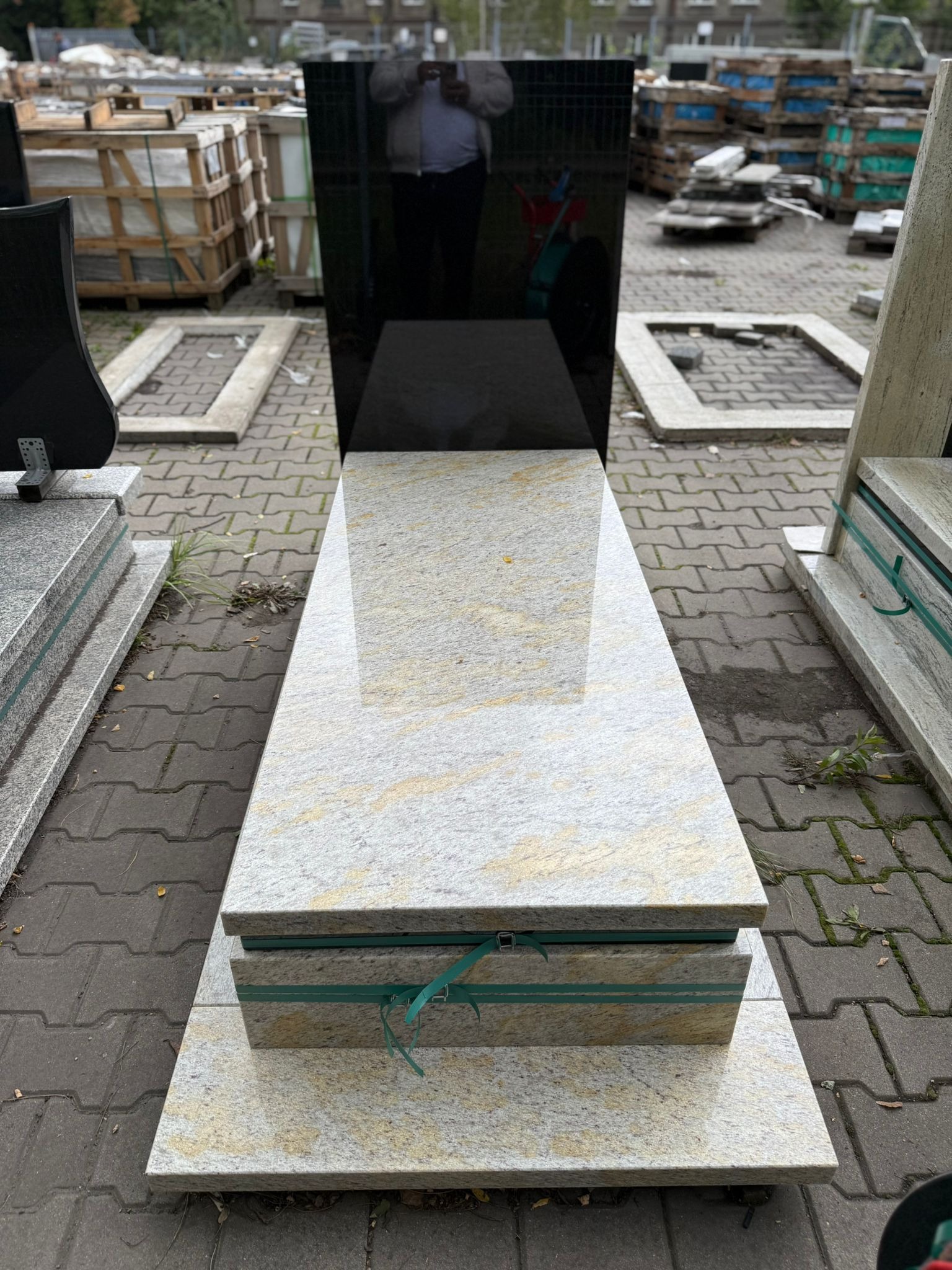 Jasny, polerowany nagrobek z czarną płytą w tle, zabezpieczony zieloną taśmą. Widoczne inne nagrobki i kostka brukowa.