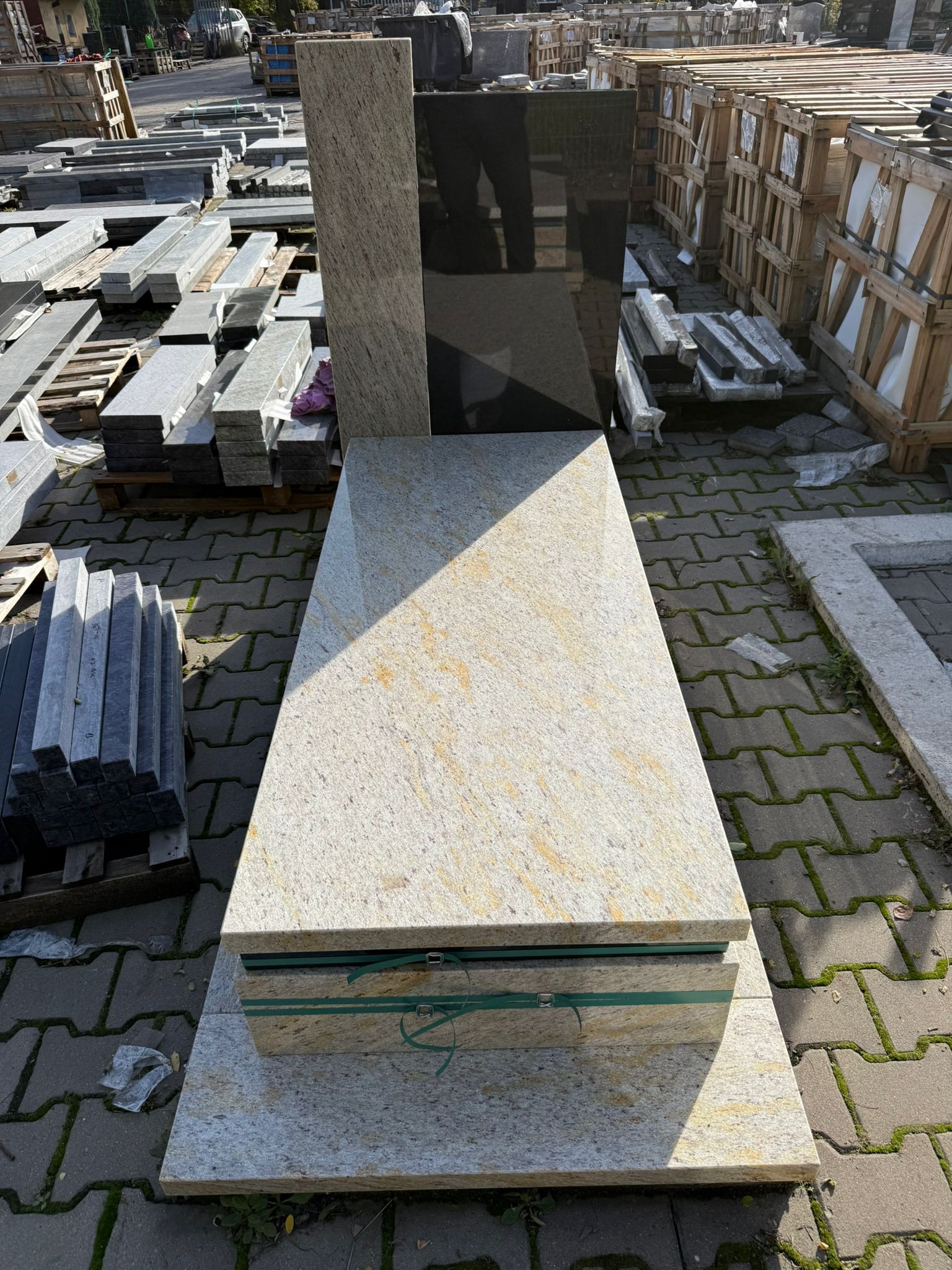 Jasny nagrobek z żółtymi żyłkami, zabezpieczony taśmami, na tle magazynu kamieniarskiego z paletami i elementami nagrobków.