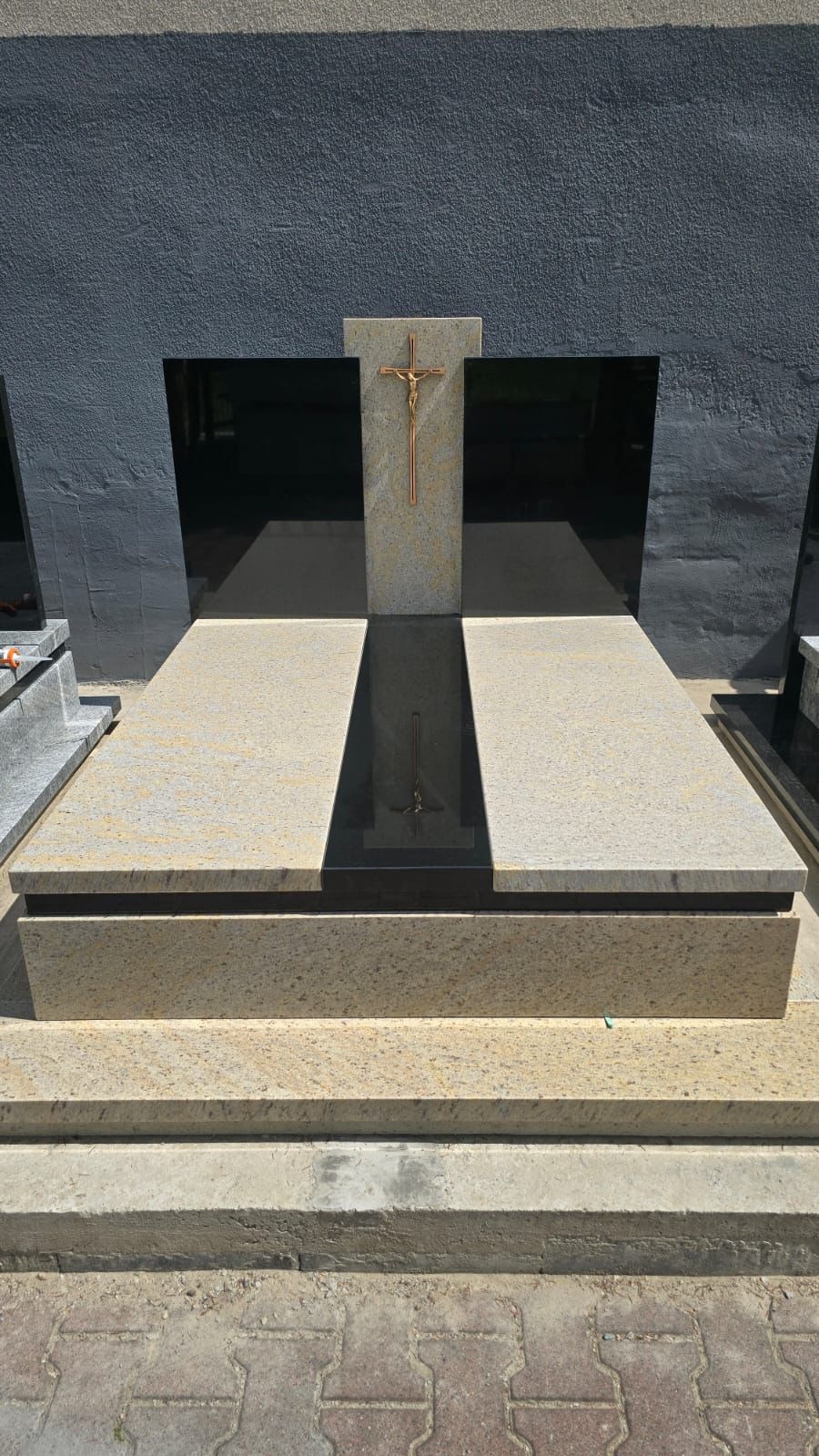 Jasny, granitowy nagrobek z ciemnymi wstawkami i złotym krzyżem, ustawiony na betonowym podłożu. Widok z dołu, oświetlenie dzienne.