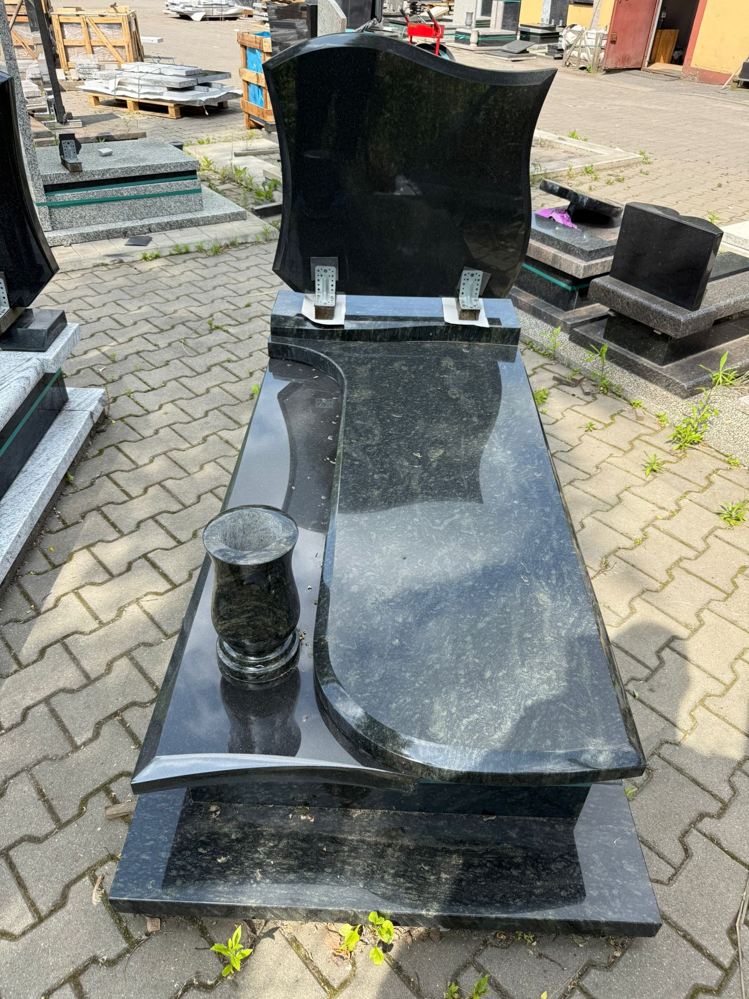 Czarny, polerowany nagrobek z wazonem na cmentarzu, widok z góry. Kamienna konstrukcja na tle szarej kostki brukowej i innych nagrobków.
