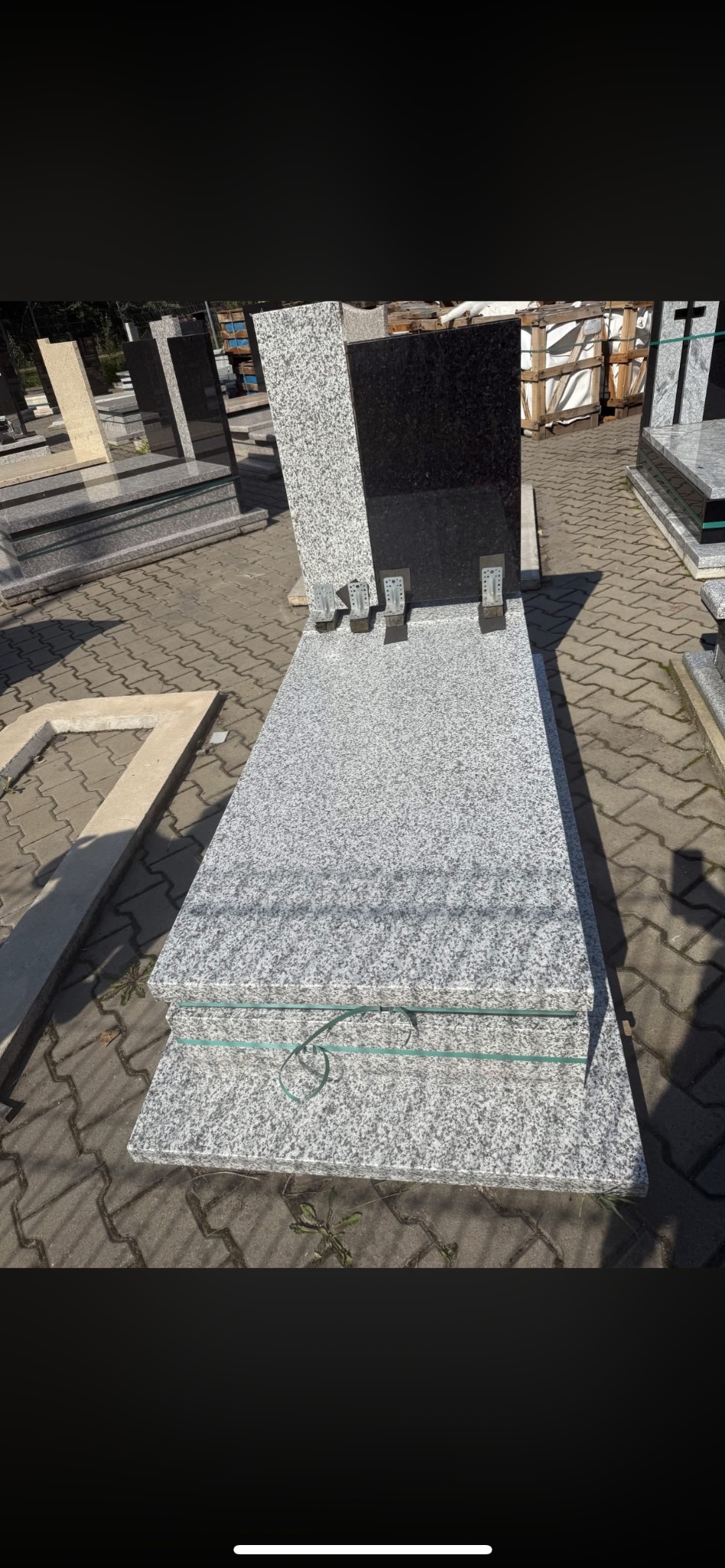 Nowoczesny, jasnoszary nagrobek z czarnym akcentem, ustawiony na kostce brukowej. Widoczne metalowe elementy montażowe i inne nagrobki w tle.