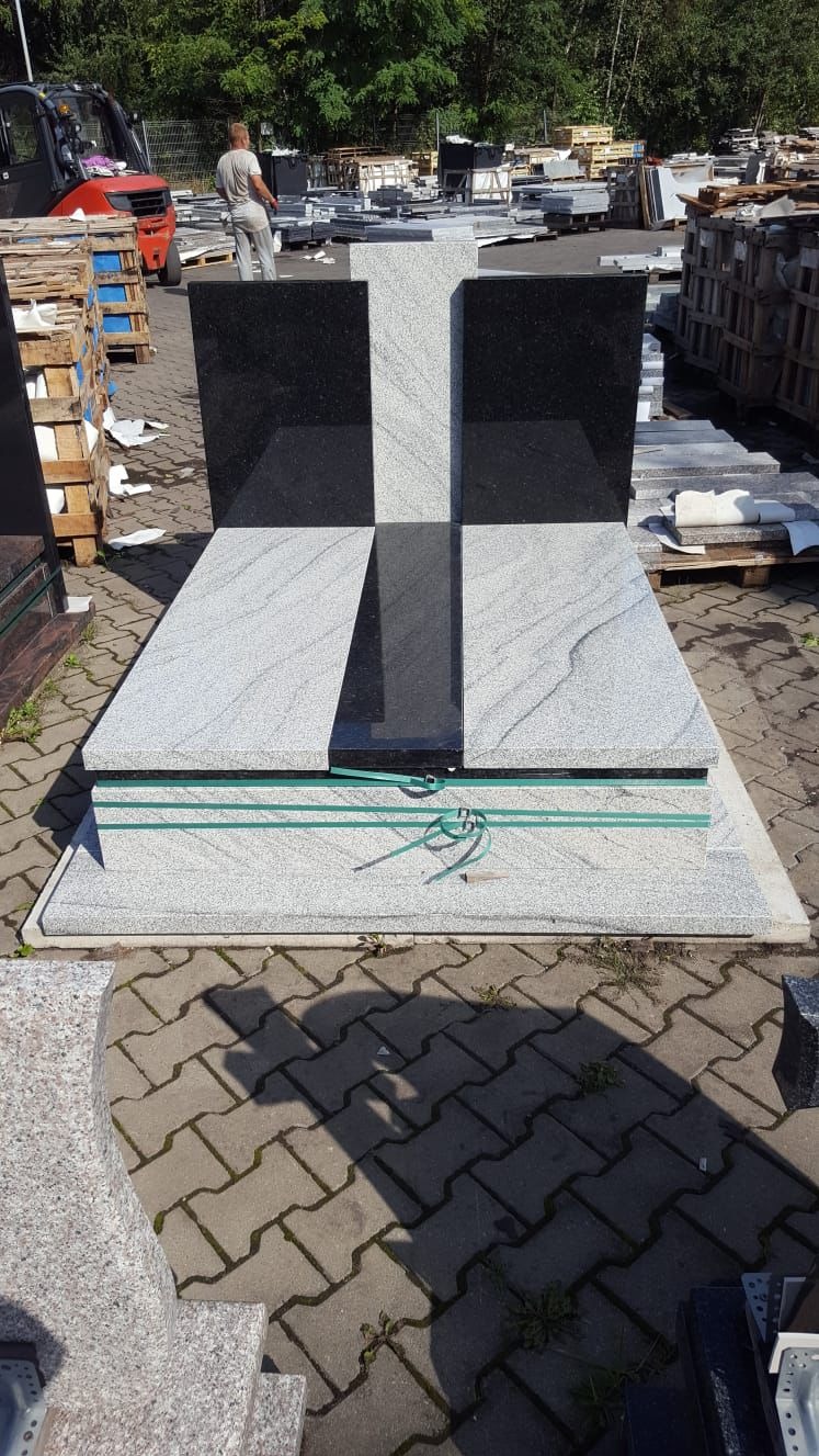 Nowy, dwukolorowy nagrobek z krzyżem, zabezpieczony zieloną taśmą, na placu kamieniarskim z widocznym wózkiem widłowym i osobą w tle.