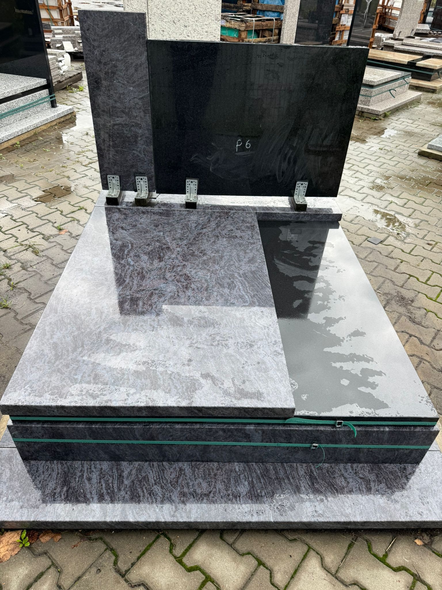 Nowy, dwukolorowy nagrobek z szarego i czarnego kamienia, z widocznymi metalowymi elementami mocującymi, ustawiony na zewnątrz na mokrej kostce brukowej.
