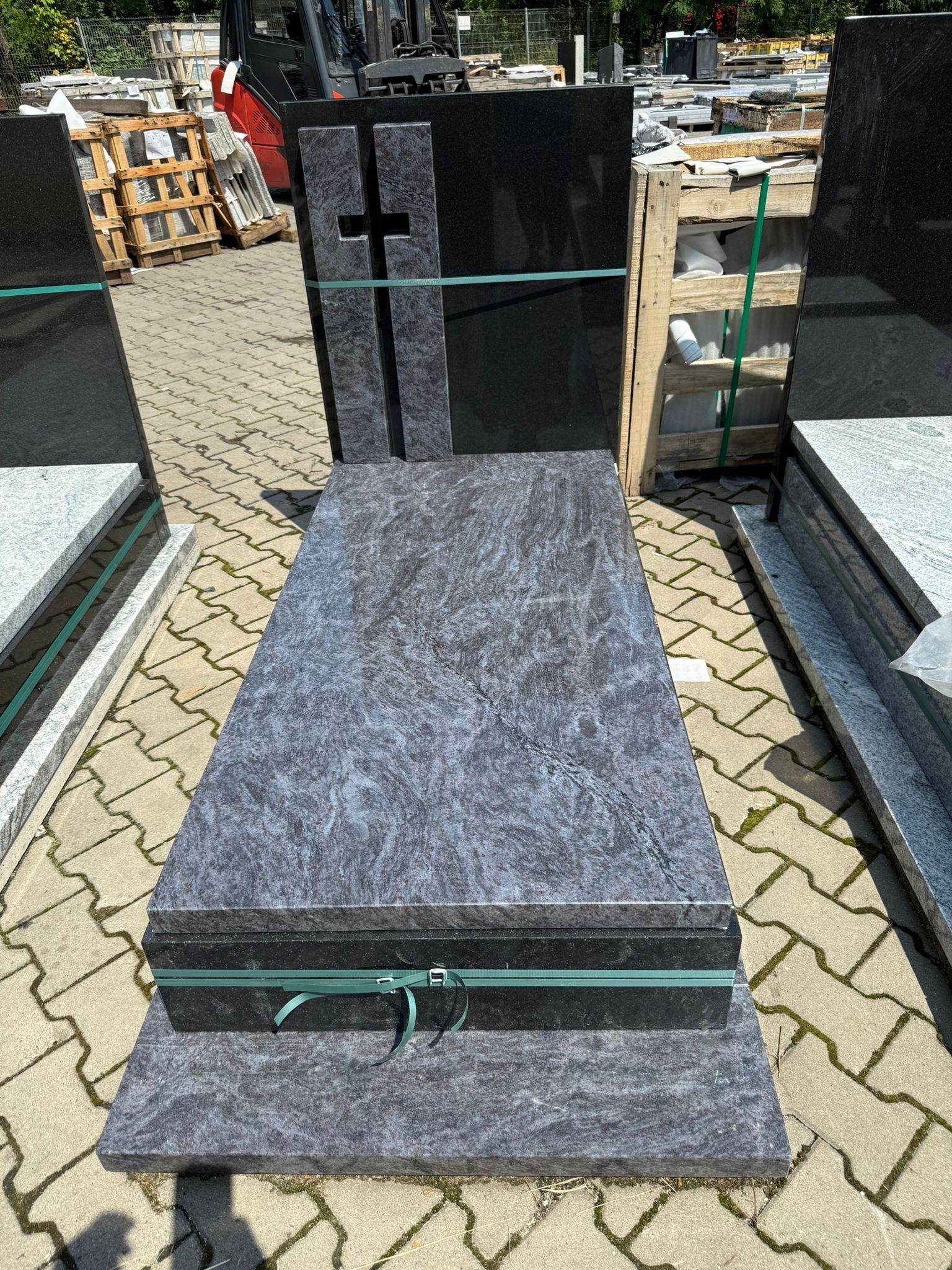 Granitowy nagrobek w odcieniach szarości z krzyżem, zabezpieczony zieloną taśmą, na tle kostki brukowej i palet. Widoczny wózek widłowy w tle.