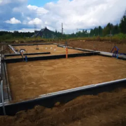 Przygotowany teren pod budowę domu jednorodzinnego: wykopane fundamenty z izolacją, zasypane piaskiem, z wystającymi rurami instalacyjnymi, w tle widoczny dom i zalesiony teren.