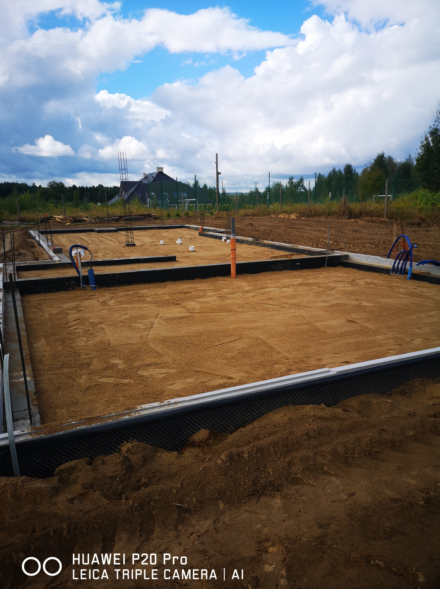 Przygotowany teren pod budowę domu jednorodzinnego: wykopane fundamenty z izolacją, zasypane piaskiem, z wystającymi rurami instalacyjnymi, w tle widoczny dom i zalesiony teren.