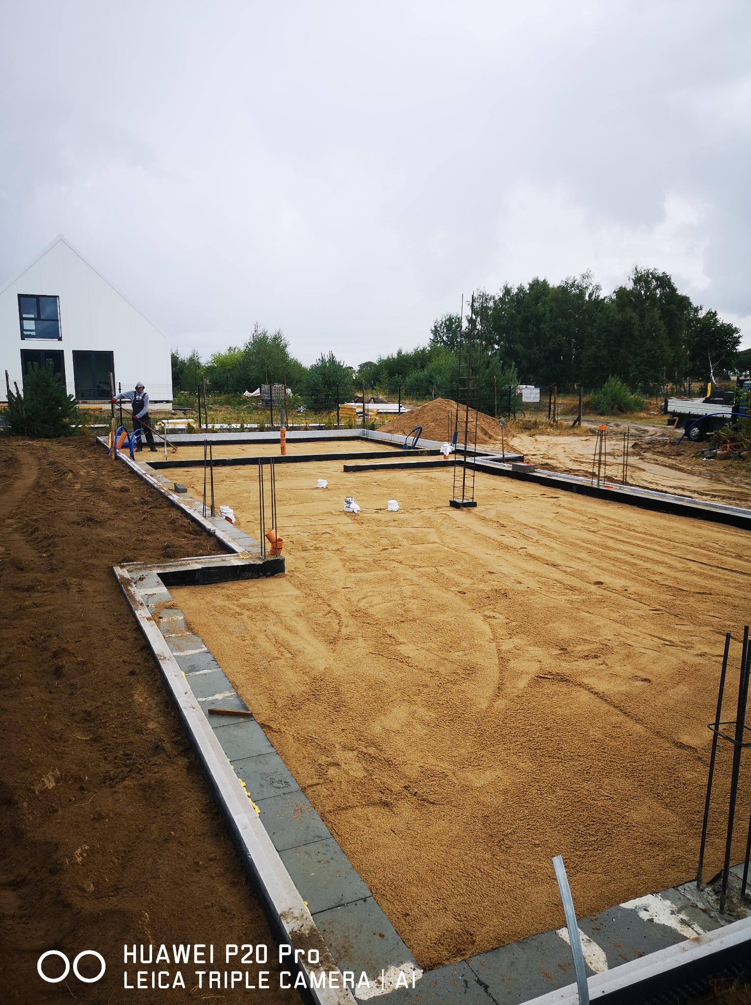 Przygotowanie terenu pod budowę domu jednorodzinnego: wyrównany piasek wewnątrz zbrojonych fundamentów, pracownik z narzędziem w tle, nowoczesny biały dom w tle, pochmurne niebo.