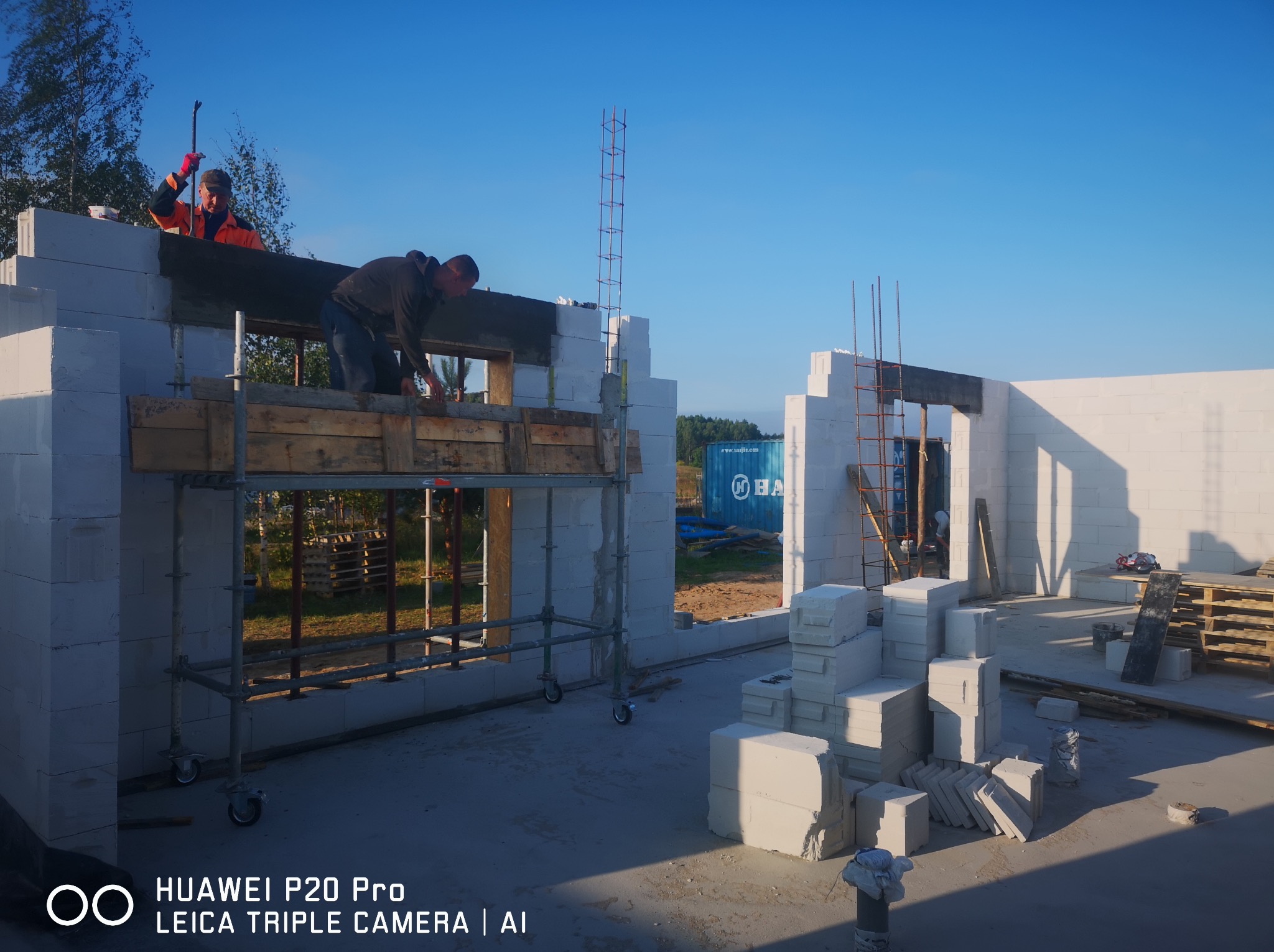 Wznoszenie ścian domu z białych bloczków, widoczni dwaj pracownicy budowlani na rusztowaniu, jeden z łomem w ręku, w tle błękitny kontener i fragment lasu.