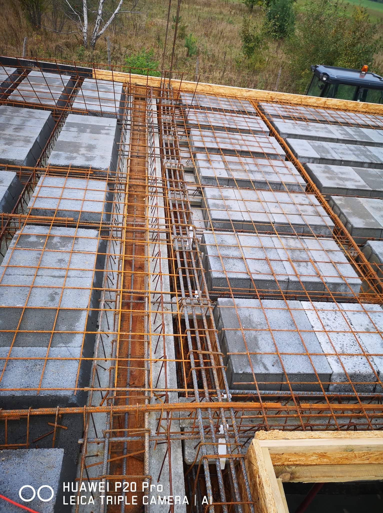 Wieniec stropowy z szalunkiem i zbrojeniem, widoczne bloczki betonowe i wystające pręty zbrojeniowe, w tle zarys traktora.