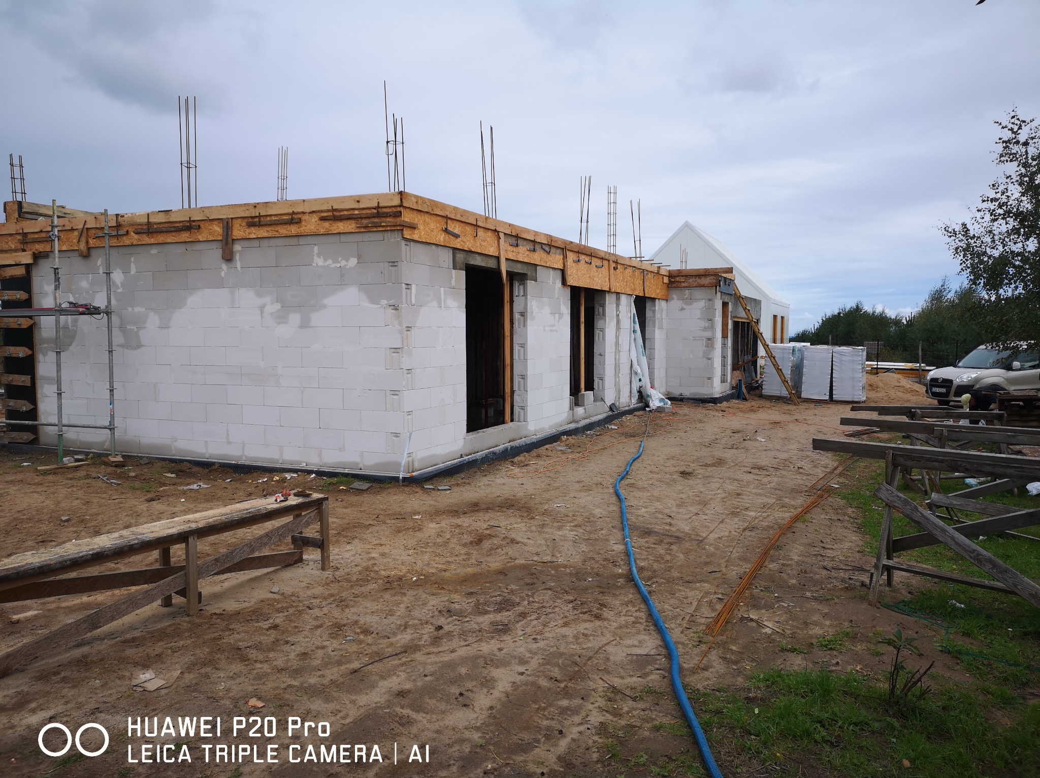 Budowa domu z białych bloczków, widoczne zbrojenia, drewniane szalunki i materiały budowlane na placu budowy w pochmurny dzień.