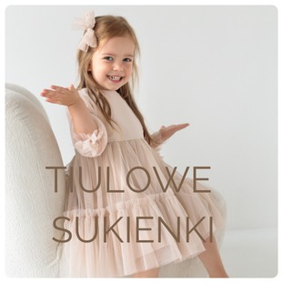 Uśmiechnięta dziewczynka w tiulowej sukience w kolorze pudrowego różu, siedząca na kremowym fotelu, z kokardą we włosach i rozłożonymi rękami.