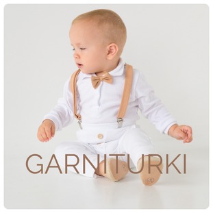Uroczy niemowlak w eleganckim białym komplecie z muchą i szelkami w odcieniach beżu, siedzący na białym tle. Widoczne małe buciki. Na zdjęciu widoczny napis 'GARNITURKI'.