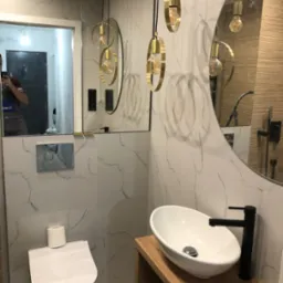 Nowoczesna łazienka z owalną umywalką nablatową na drewnianym blacie, wiszącą toaletą, okrągłym lustrem i złotymi lampami wiszącymi na tle marmurowych płytek.