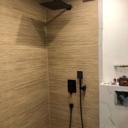 Mateusz lasok - Kabina prysznicowa z brązowymi płytkami imitującymi drewno, czarna deszczownica i bateria, białe płytki imitujące marmur w sąsiedztwie, widoczny fragment instalacji wodnej.