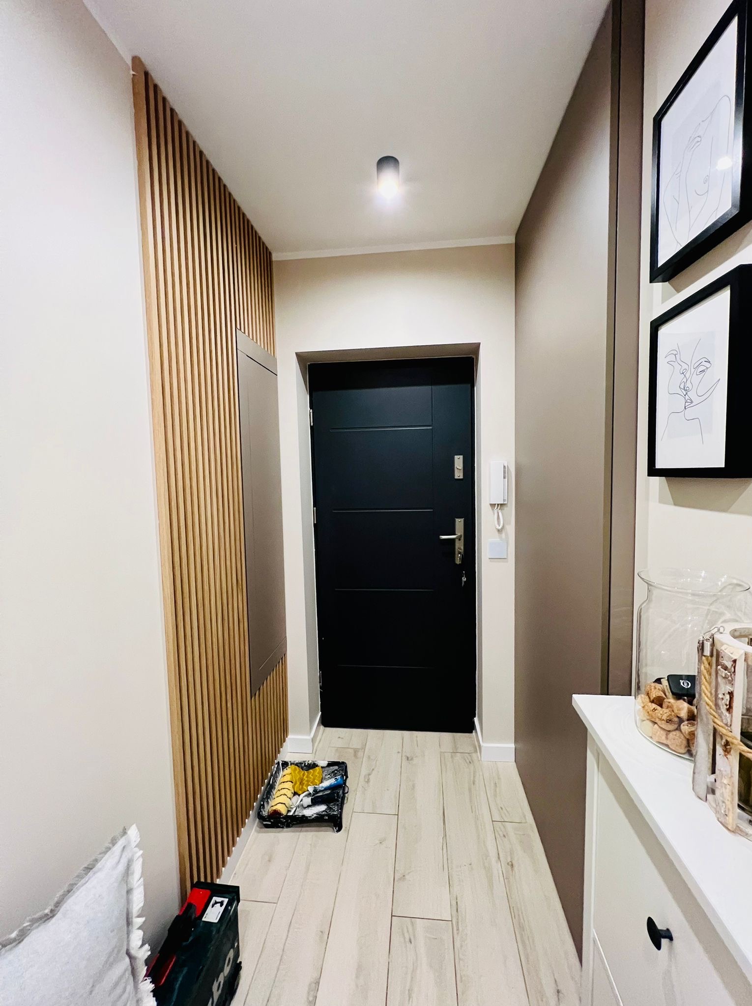 Nowoczesny przedpokój z drewnianą ścianą z lameli, czarnymi drzwiami wejściowymi i akcesoriami malarskimi w rogu
