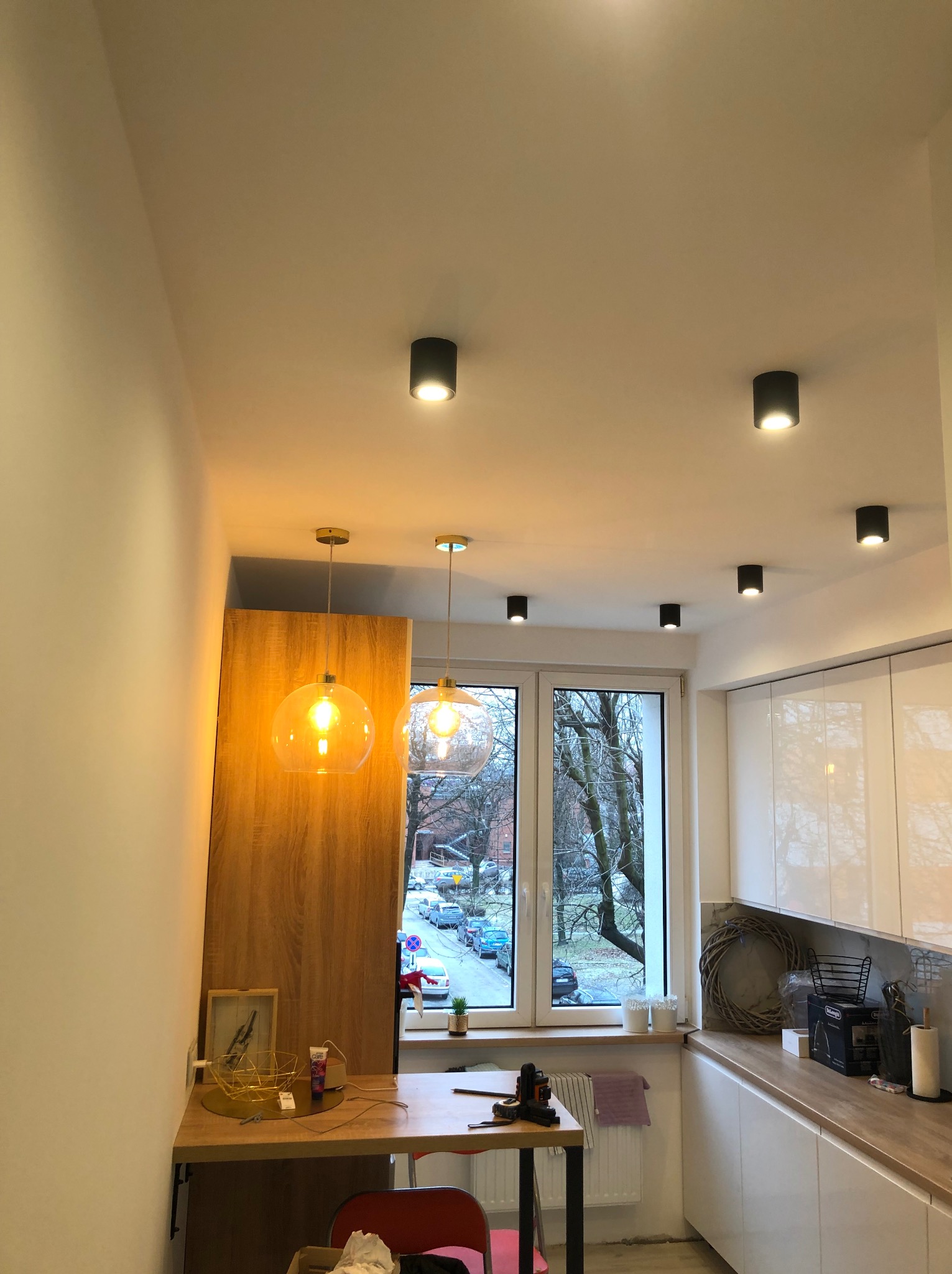 Nowoczesna kuchnia z białymi szafkami, drewnianym blatem i okrągłymi lampami wiszącymi z żarówkami Edisona, oświetlona również czarnymi lampami sufitowymi punktowymi, widok na ulicę przez okno.