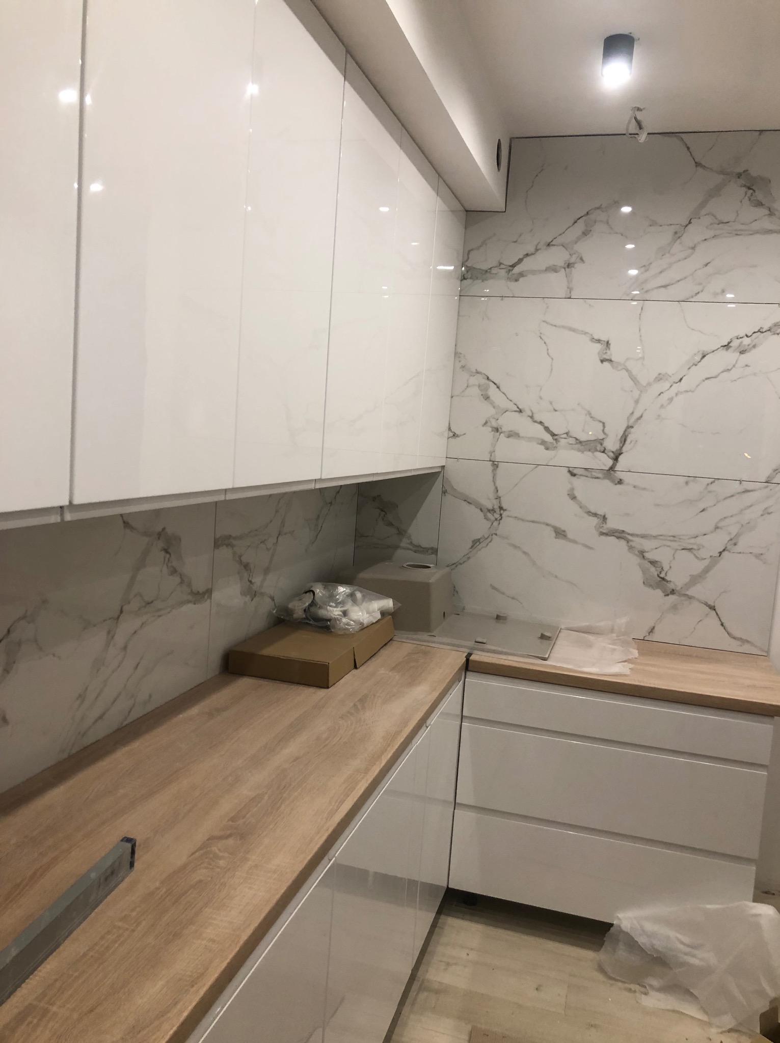 Fragment kuchni w trakcie wykańczania: białe, lakierowane szafki wiszące i dolne, drewnopodobny blat, marmuropodobne płytki na ścianie, widoczne narzędzia i materiały budowlane.