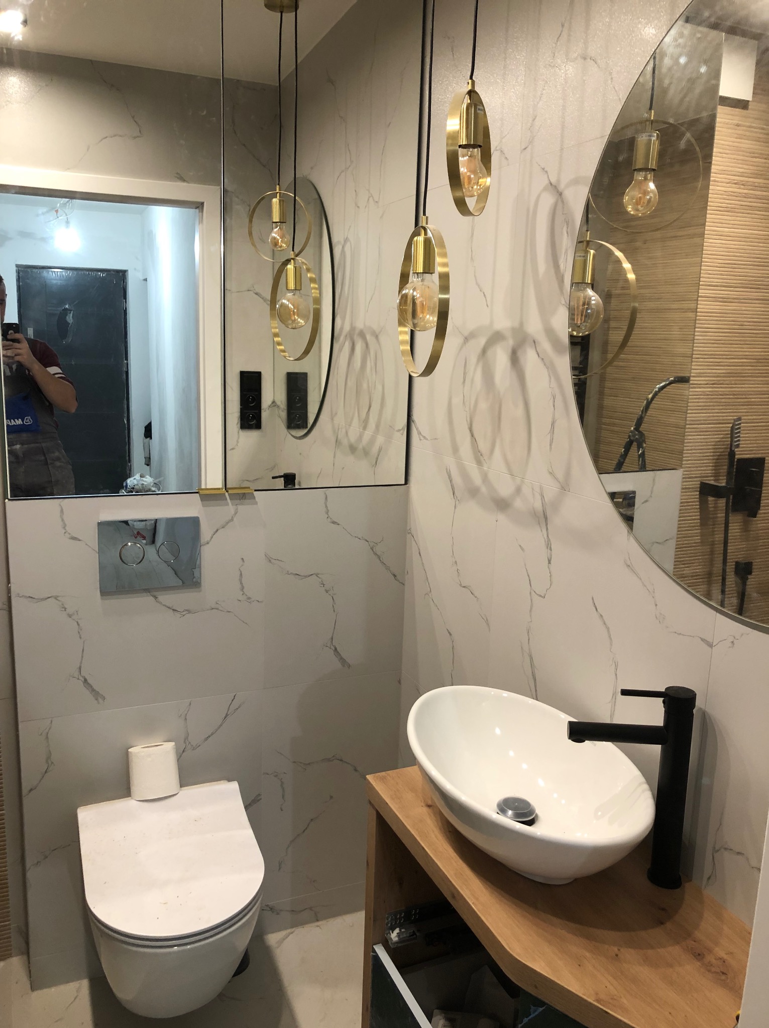 Nowoczesna łazienka z owalną umywalką nablatową na drewnianym blacie, wiszącą toaletą, okrągłym lustrem i złotymi lampami wiszącymi na tle marmurowych płytek.
