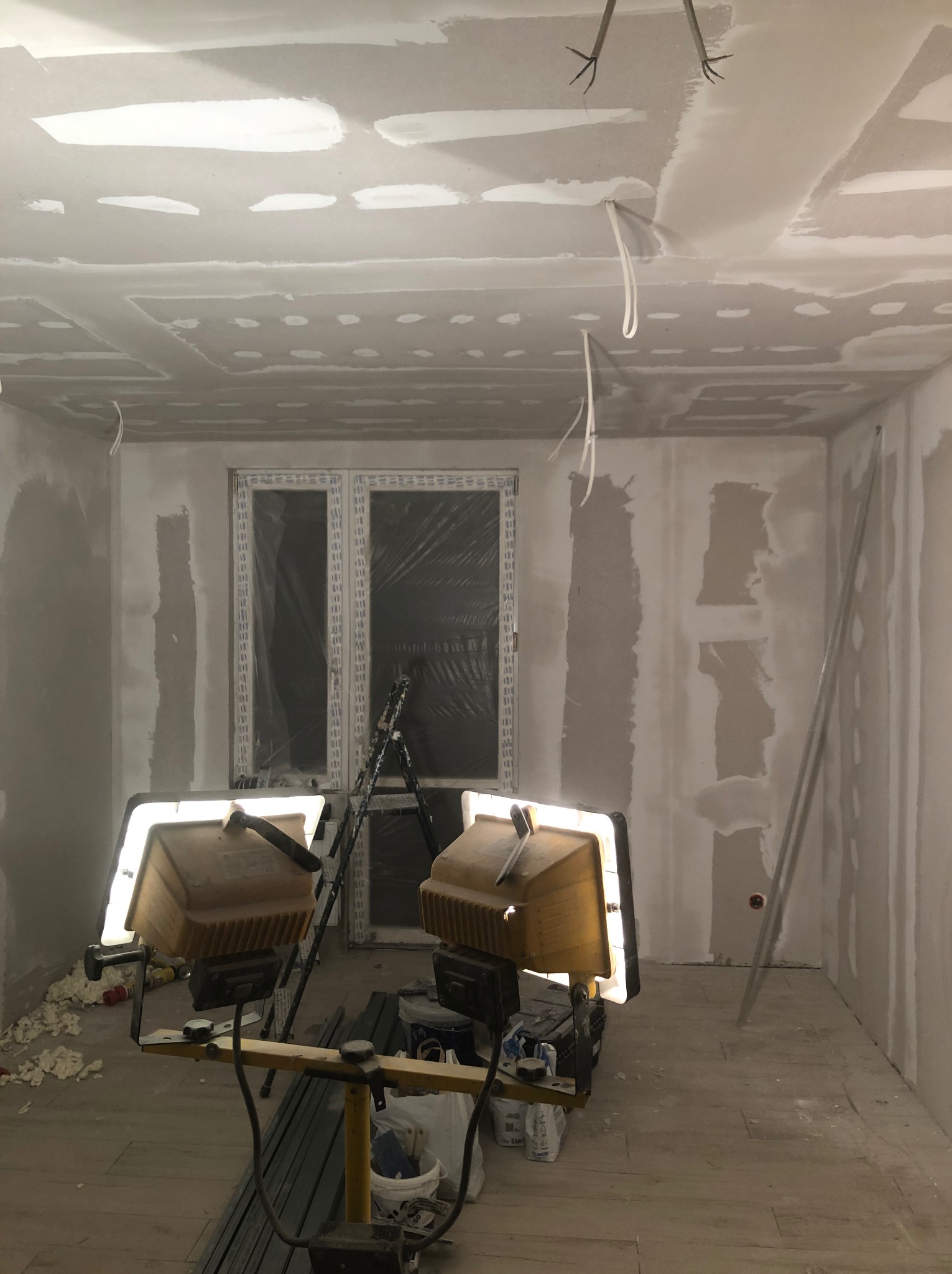 Wnętrze w trakcie prac wykończeniowych: ściany z płyt gipsowo-kartonowych z widocznymi łączeniami szpachlowanymi, okno zabezpieczone folią, rozstawione lampy budowlane i drabina, podłoga częściowo...