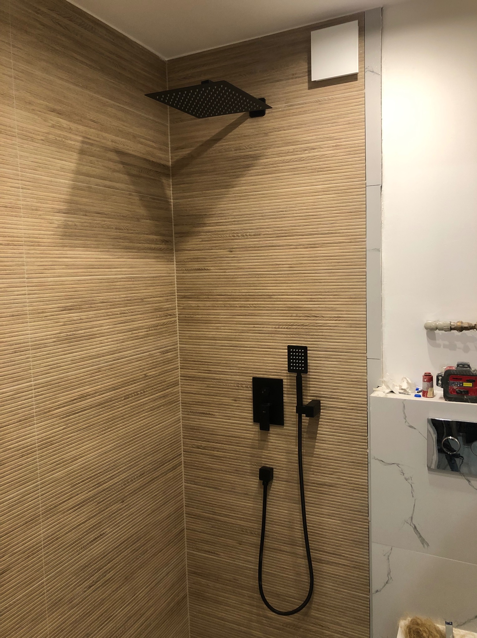 Kabina prysznicowa z brązowymi płytkami imitującymi drewno, czarna deszczownica i bateria, białe płytki imitujące marmur w sąsiedztwie, widoczny fragment instalacji wodnej.