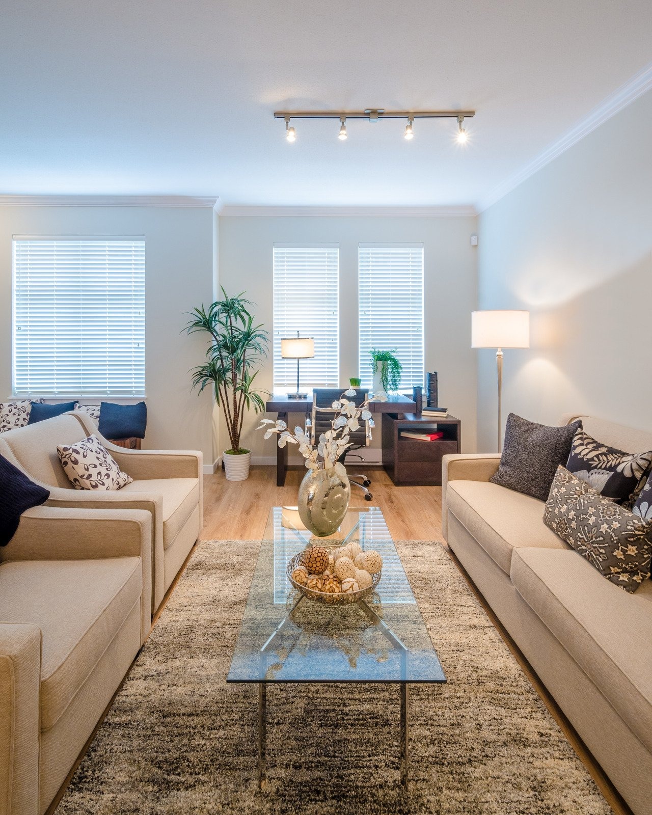 Neutralny salon z dwoma beżowymi sofami ustawionymi naprzeciwko siebie, szklanym stolikiem kawowym z dekoracyjną misą, drewnianą podłogą i dywanem w odcieniach szarości, oświetlony listwą...