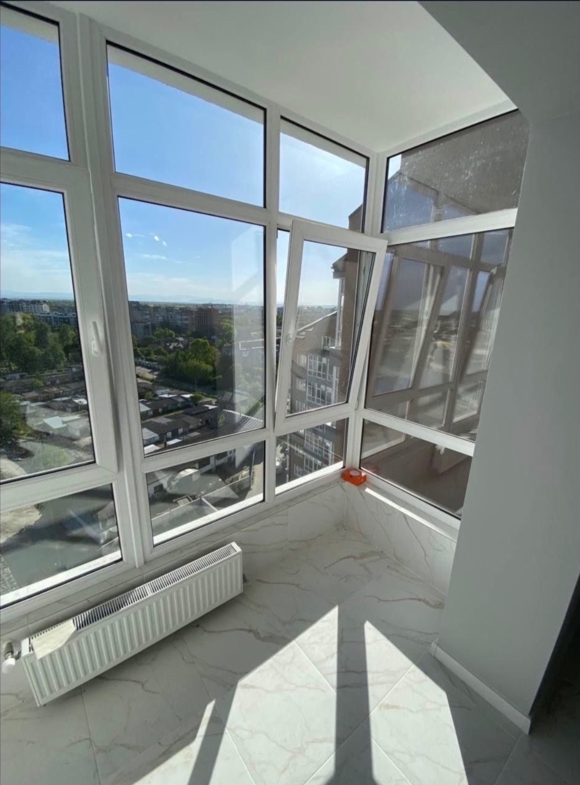 Zabudowany balkon z białą ramą okienną, marmurową podłogą i grzejnikiem, widok na miasto w słoneczny dzień.