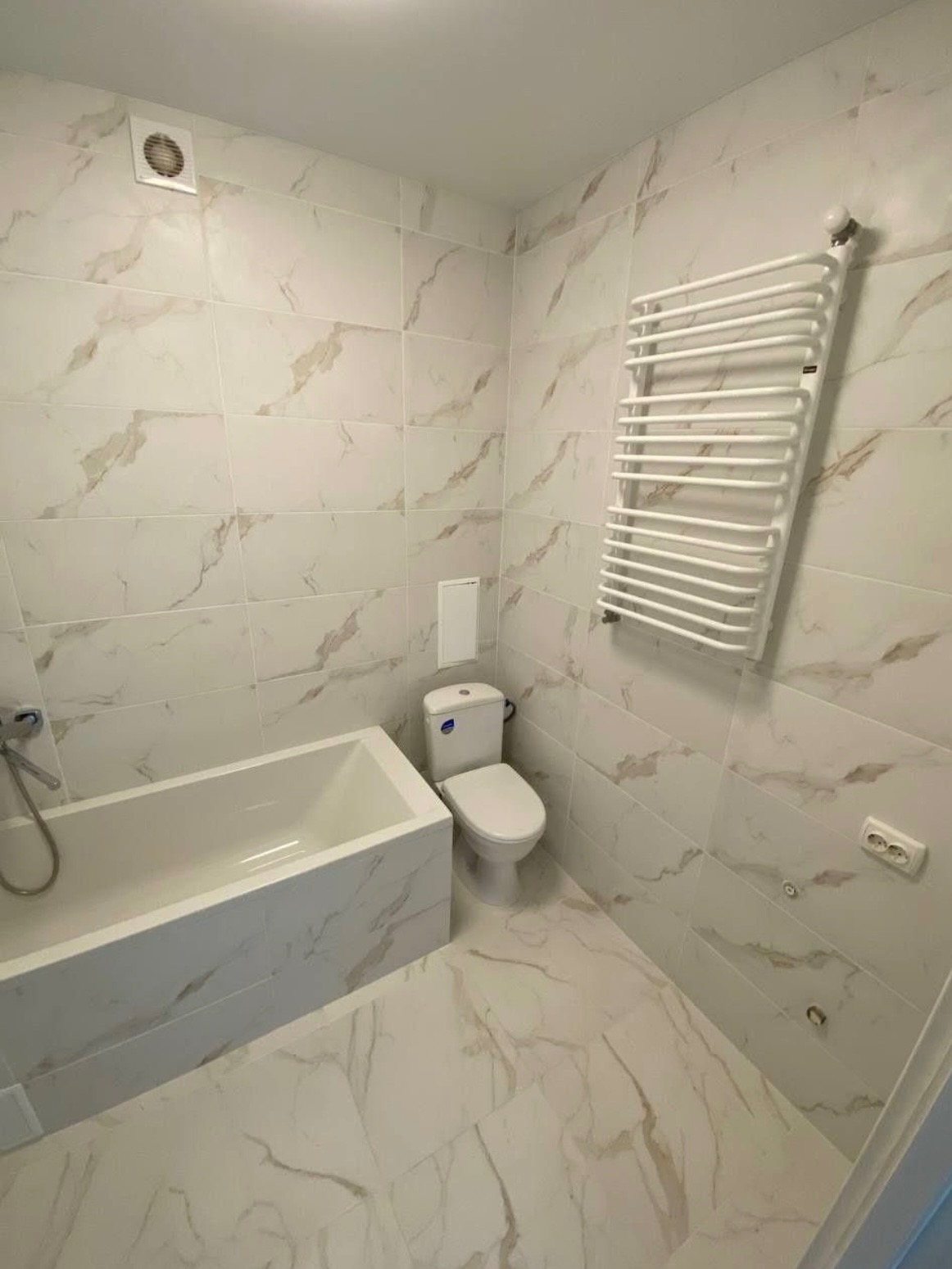 Wykończona łazienka z białymi płytkami imitującymi marmur, z wanną zabudowaną płytkami, toaletą kompaktową i grzejnikiem drabinkowym na ścianie.
