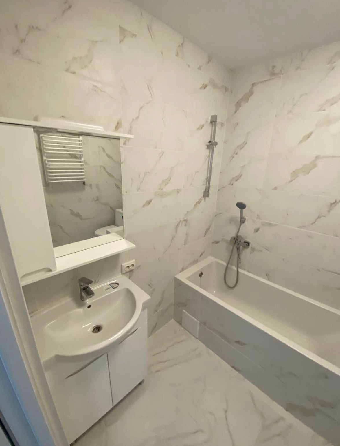 Wykończona łazienka z białymi płytkami imitującymi marmur, białą umywalką z szafką, wanną i grzejnikiem drabinkowym widocznym w lustrze.