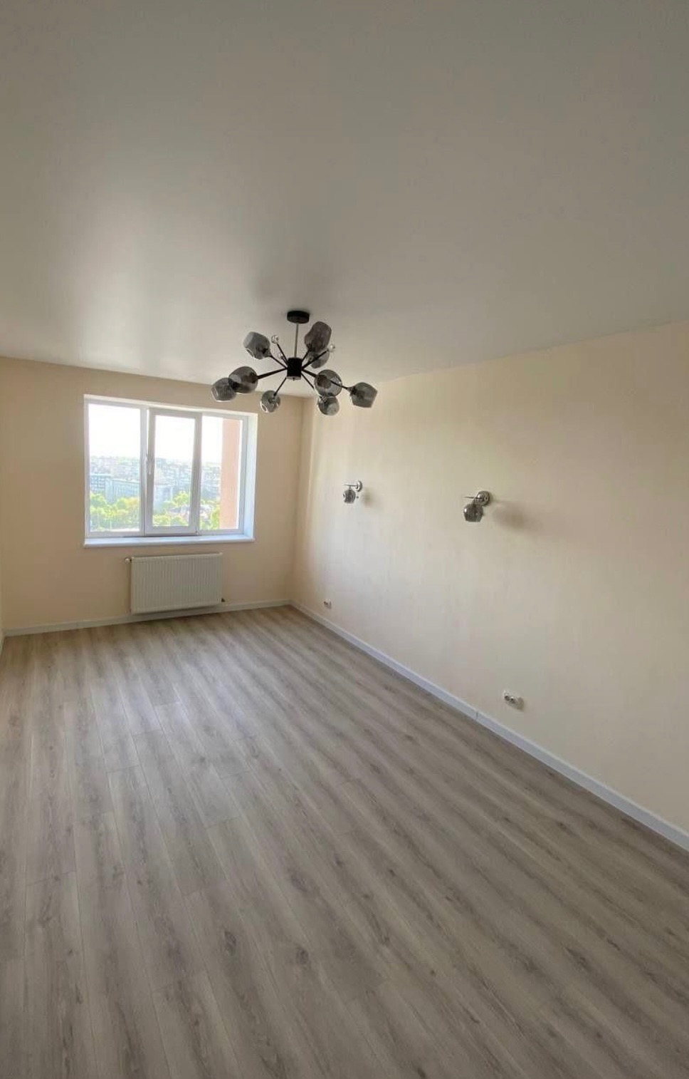 Pusty pokój po remoncie, z panelami podłogowymi w odcieniach szarości, kremowymi ścianami, nowoczesnym żyrandolem i dwoma kinkietami, widok na miasto przez okno.