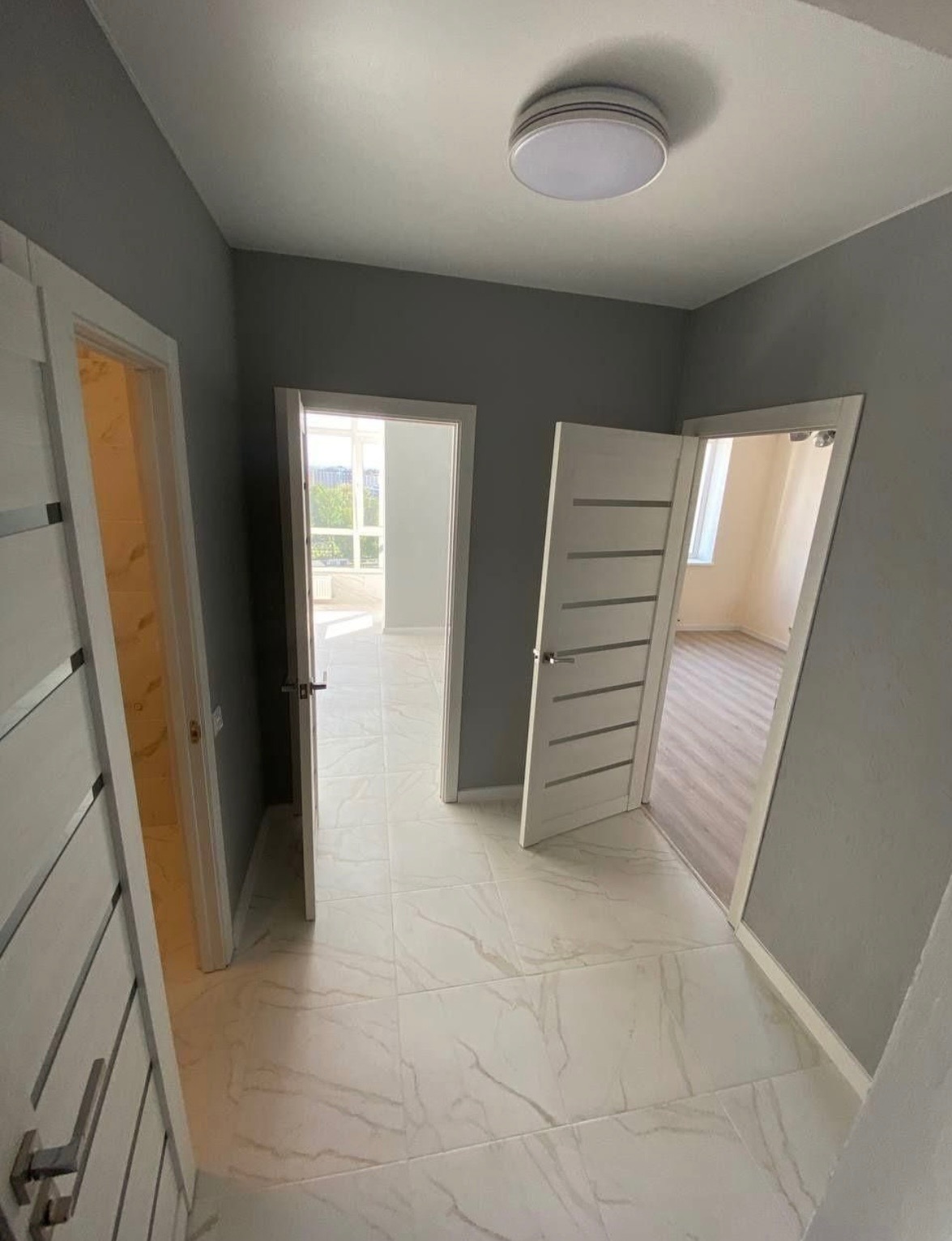 Korytarz w odcieniach szarości z trzema białymi drzwiami i białymi płytkami podłogowymi z marmurkowym wzorem.