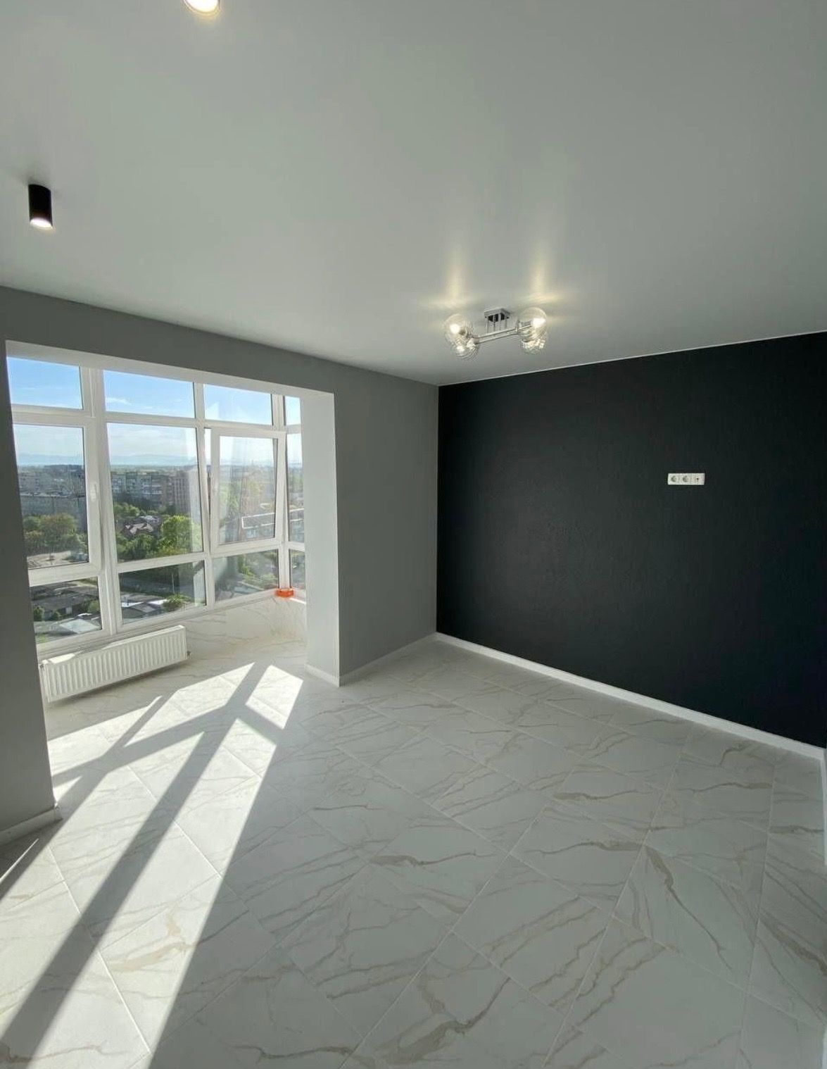 Pusty pokój po remoncie z czarną ścianą akcentującą, marmurowe płytki podłogowe w odcieniach bieli i szarości, duże okno z widokiem na miasto i silne promienie słoneczne wpadające do wnętrza.