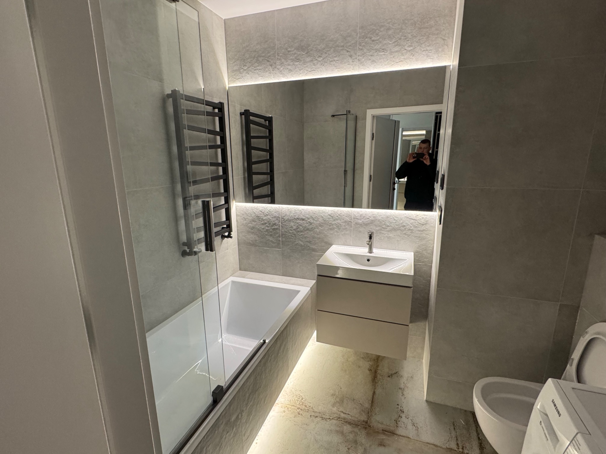 Wykończona łazienka w Warszawie z szarymi płytkami imitującymi kamień, podświetlanym lustrem, grzejnikiem drabinkowym, białą wanną z parawanem nawannowym i wiszącą szafką z umywalką.