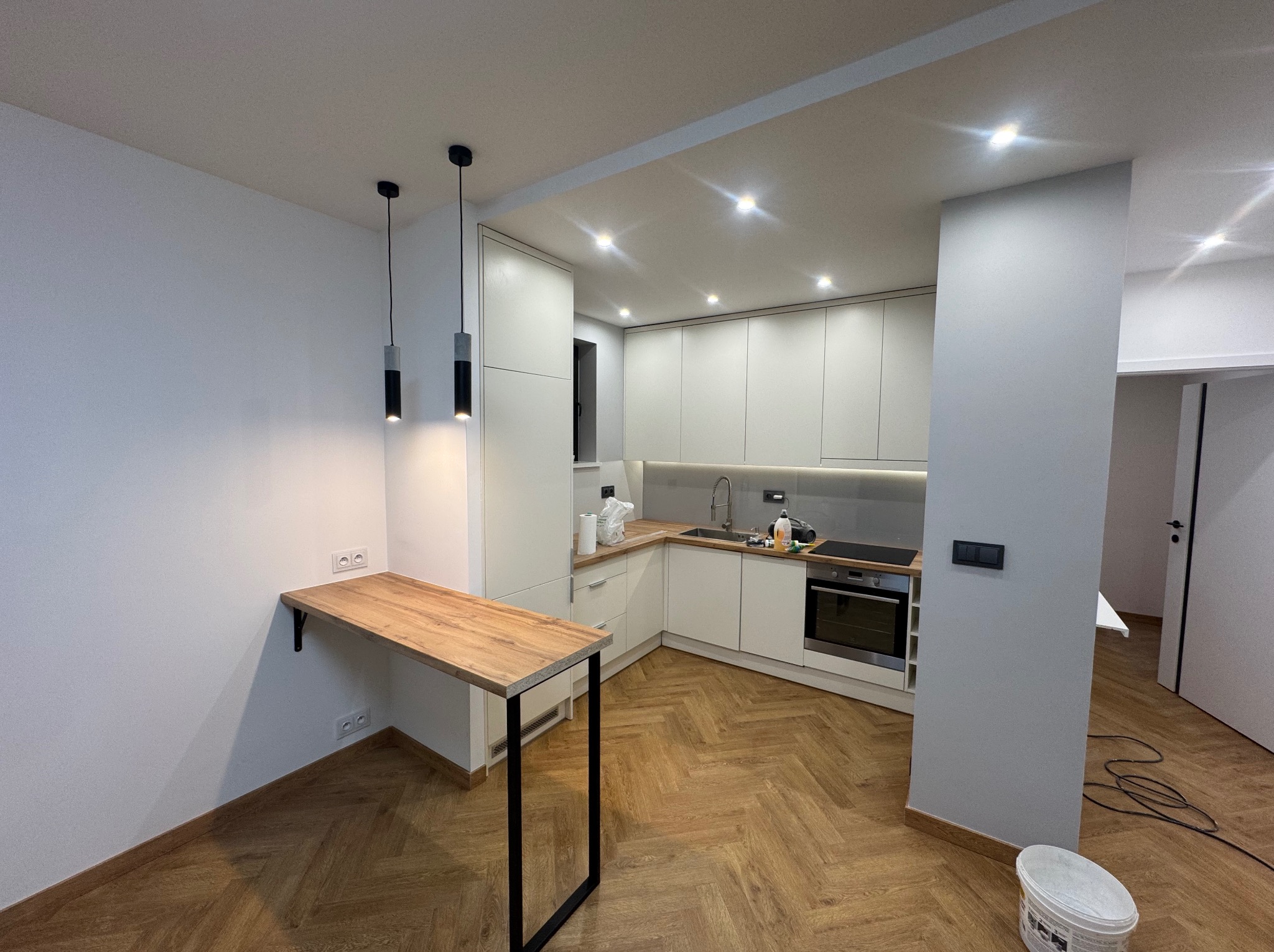 Nowoczesna kuchnia z białymi szafkami, drewnianym blatem i podłogą ułożoną w jodełkę, uzupełniona o minimalistyczne lampy wiszące i drewniany blat przymocowany do ściany.